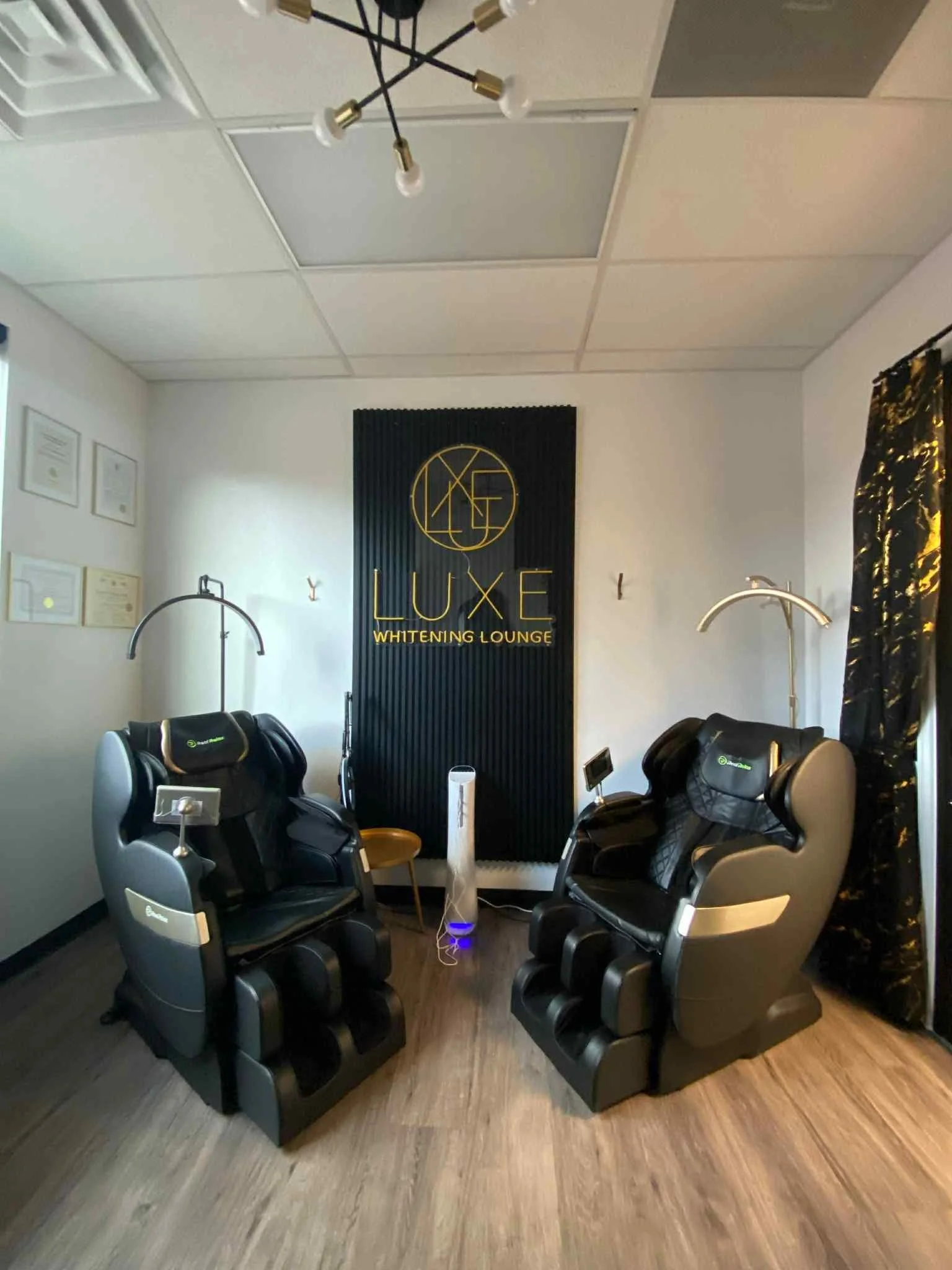 Two massage chairs in a wellness lounge with a sign reading 'Luxe Whitening Lounge' behind them, and a small fan between the chairs, in a modern treatment room.