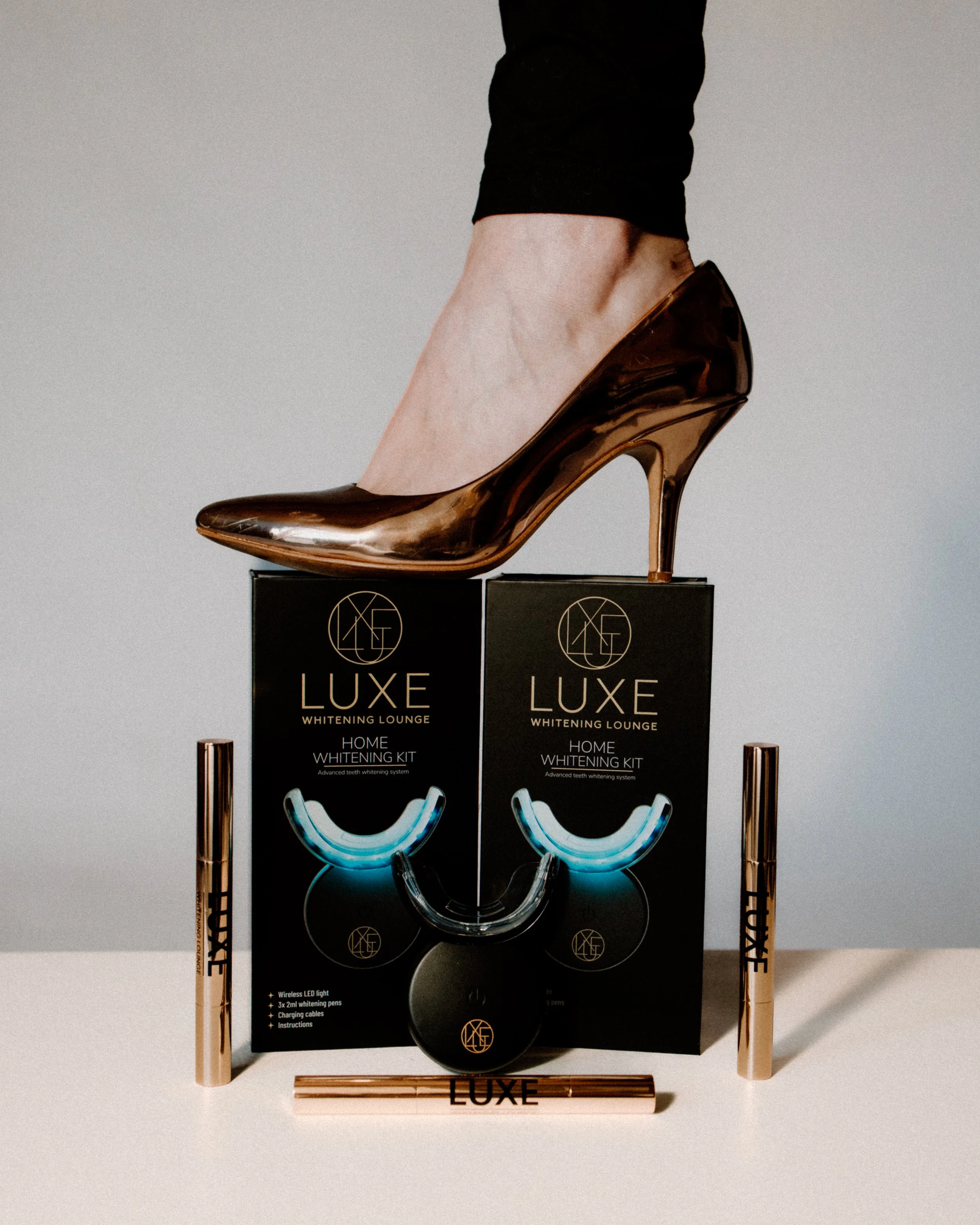 2 Black boxes labeled 'LUXE whitening kit' with a gold high-heeled shoe stepping on them, three gold-colored teeth whitening pens.