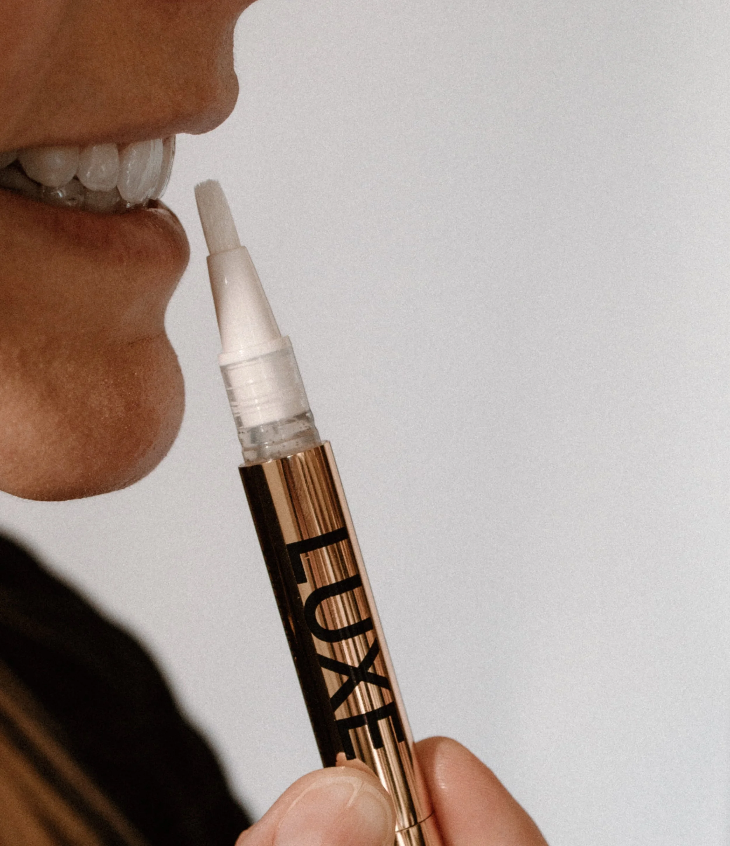 Close-up of a person holding a gold teeth whitening pen near their lips, with a section of their face and teeth visible.