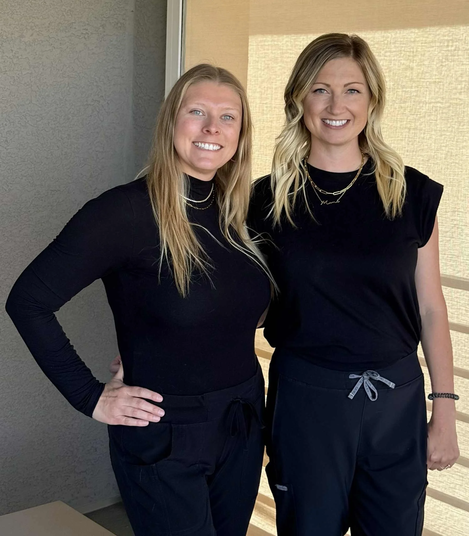 Two women wearing black clothing standing close together and smiling in front of a wall with a window and beige curtains.