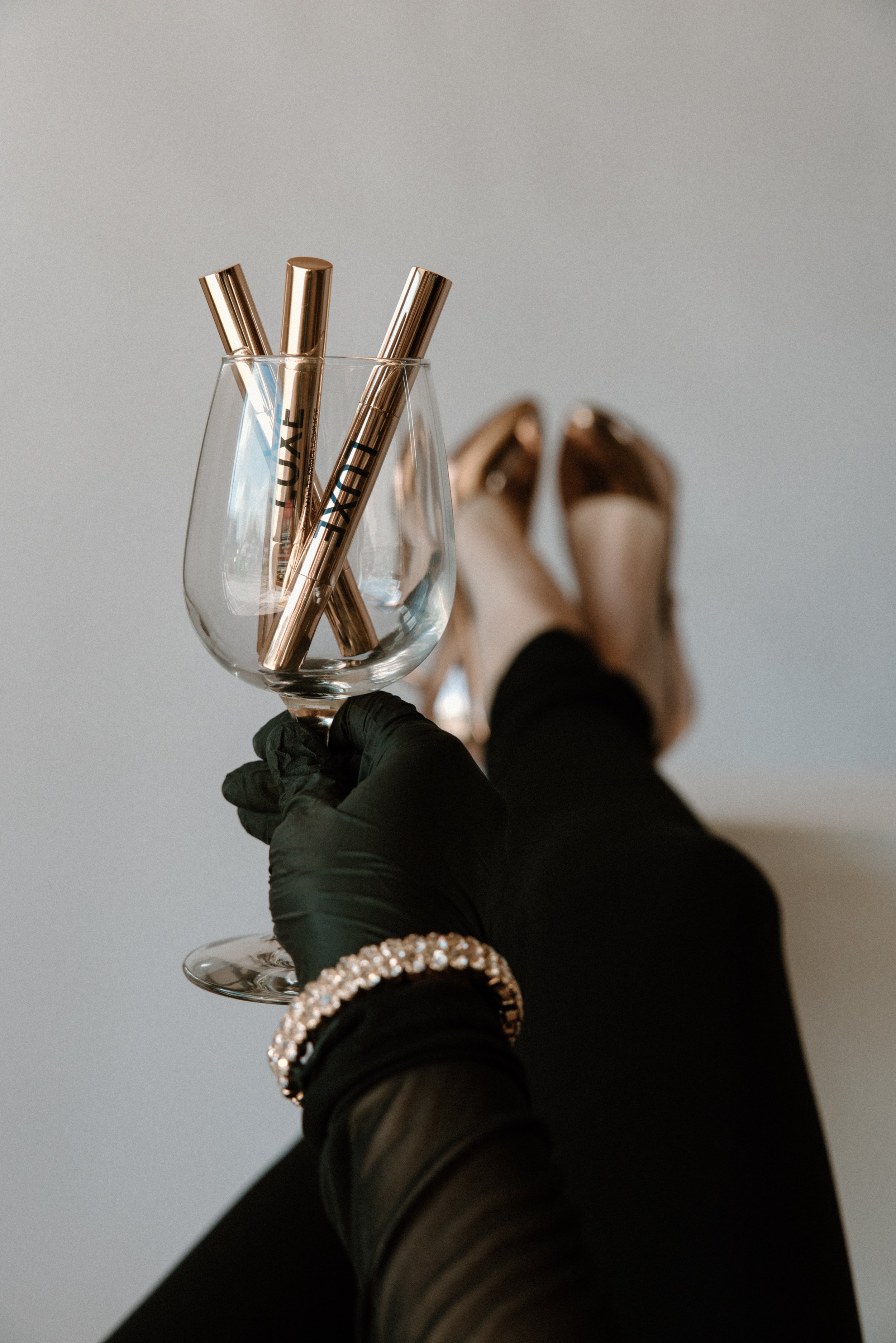 A person wearing a black glove, holding a wine glass with four metallic gold pens inside. The person is dressed in black clothing, wearing a bracelet with white stones, and rose gold shoes are visible in the background.