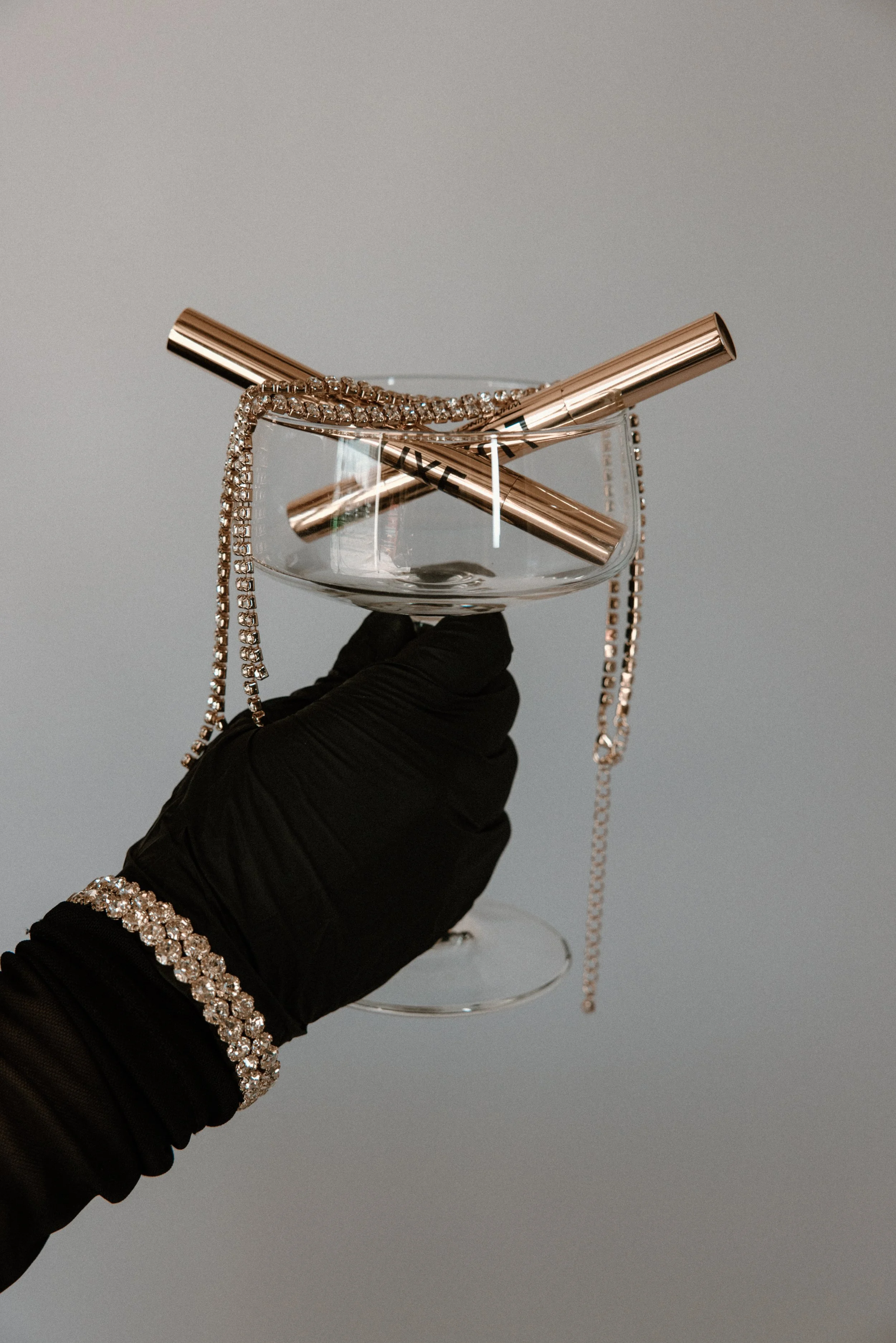 A hand wearing a black glove holding a wine glass with jewelry and metallic teeth whitening pens inside it.