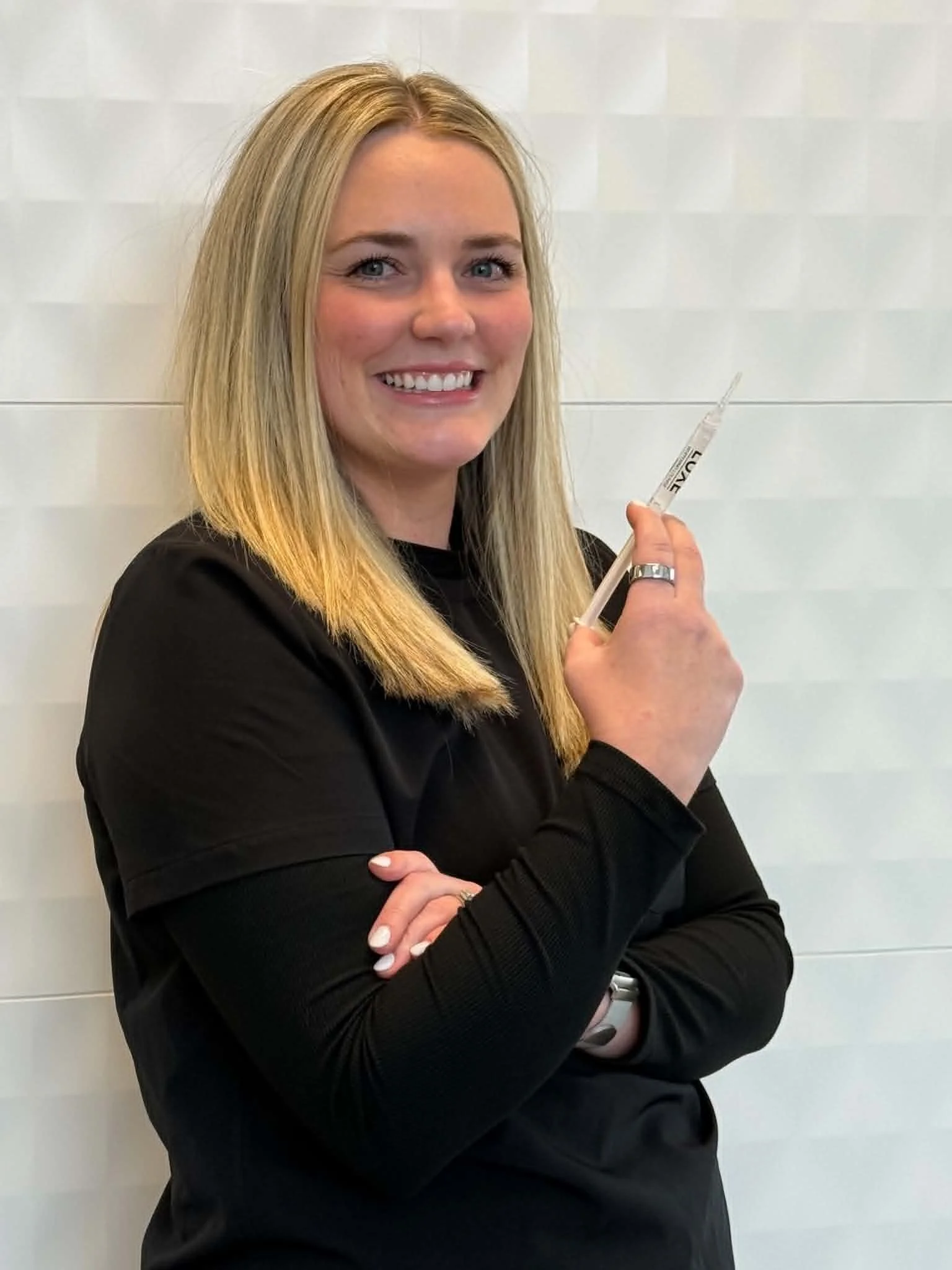 A woman with long blonde hair, wearing a black long sleeve top, smiling and holding a teeth whitening syringe in her right hand, standing against a white tiled wall background.