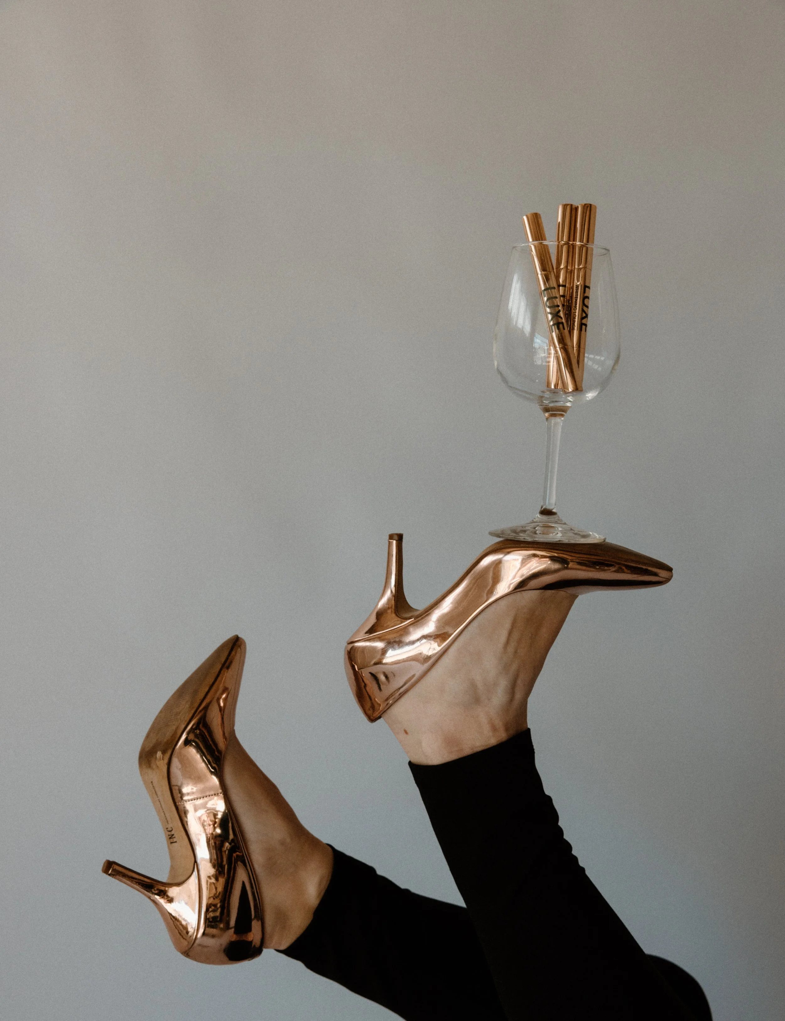 Person's leg wearing a black stocking, with a shiny rose gold high-heeled shoe and a white wine glass with rose gold straws, against a plain grey background.