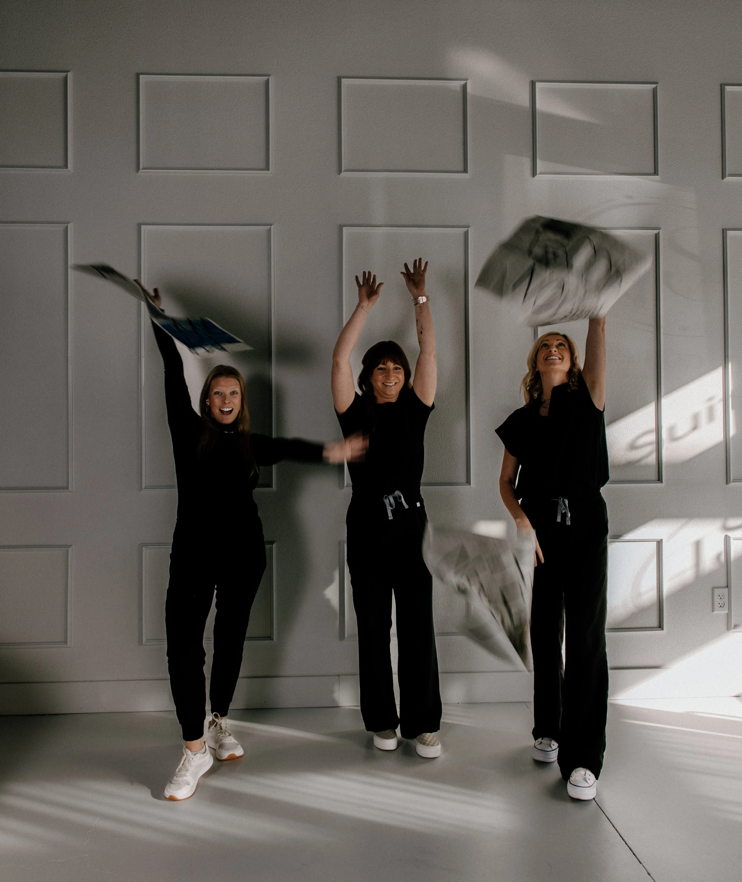 Three LUXE employees in black outfits and white shoes, happily tossing newspapers into the air in front of a white paneled wall.