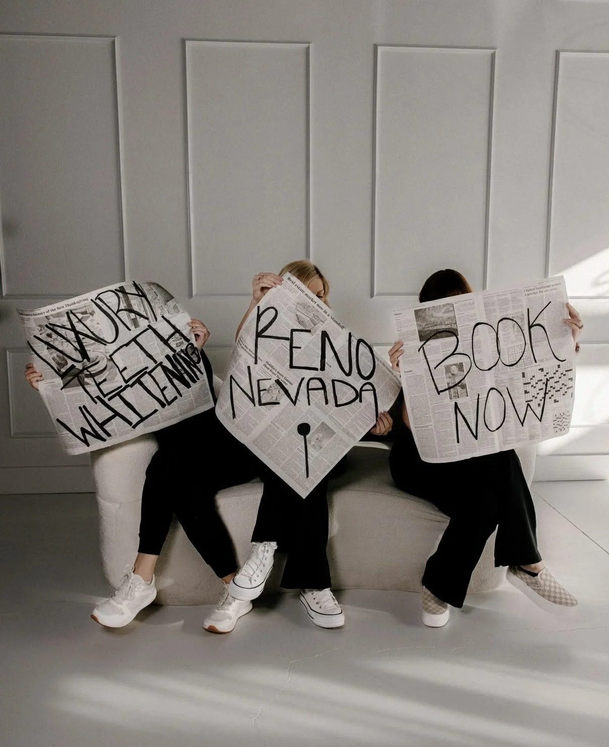 Three women holding newspaper-style signs that read “Luxury Teeth Whitening,” “Book Now,” and “Reno Nevada,” representing a modern luxury teeth whitening studio and professional whitening services.