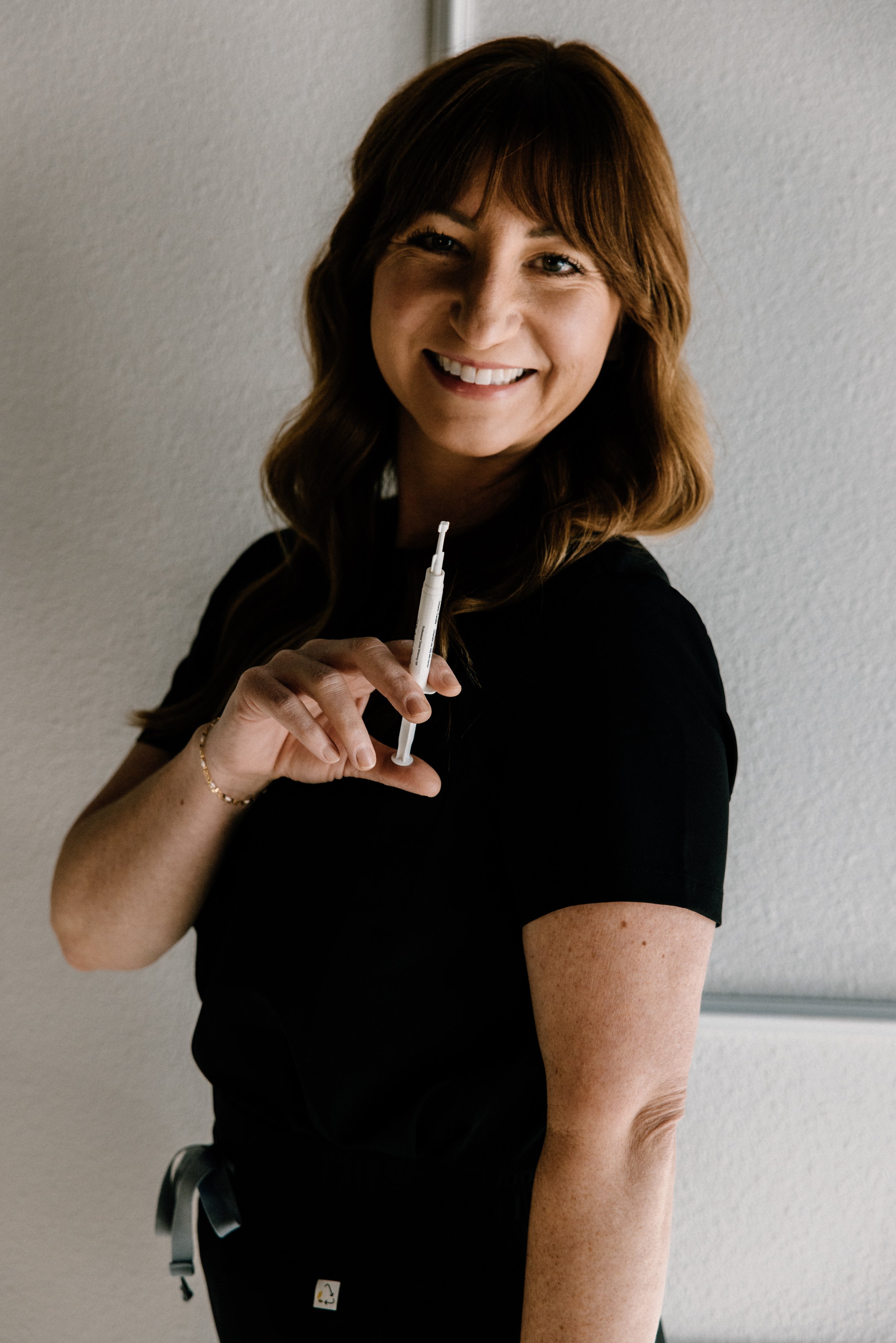 Woman with brown hair and light skin holding a teeth whitneing gel syringe, smiling, standing against a plain wall.