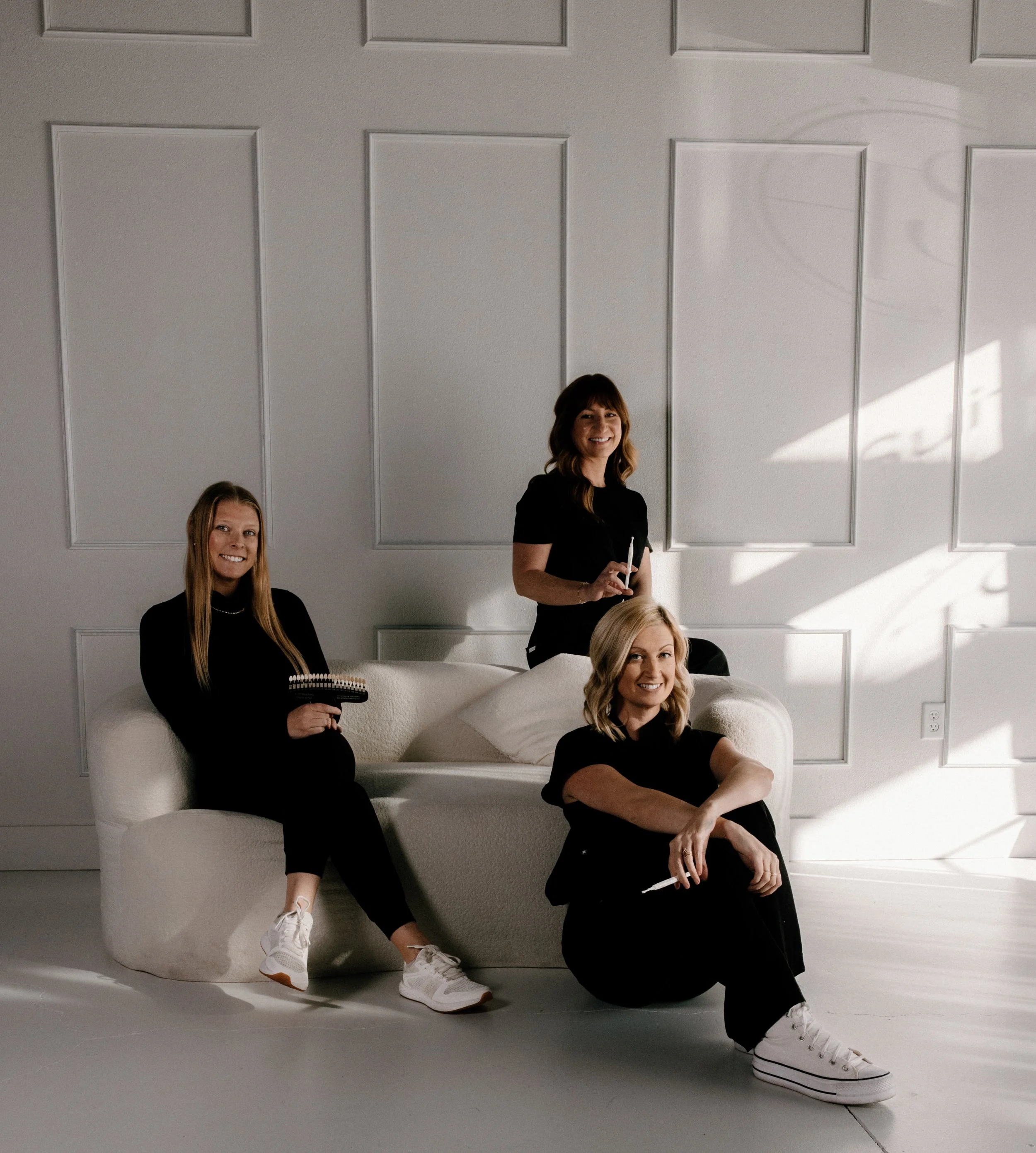 Three women with light skin in black clothing sitting and standing on and near a white sofa in a minimalist, white room with decorative wall panels and sunlight streaming in.