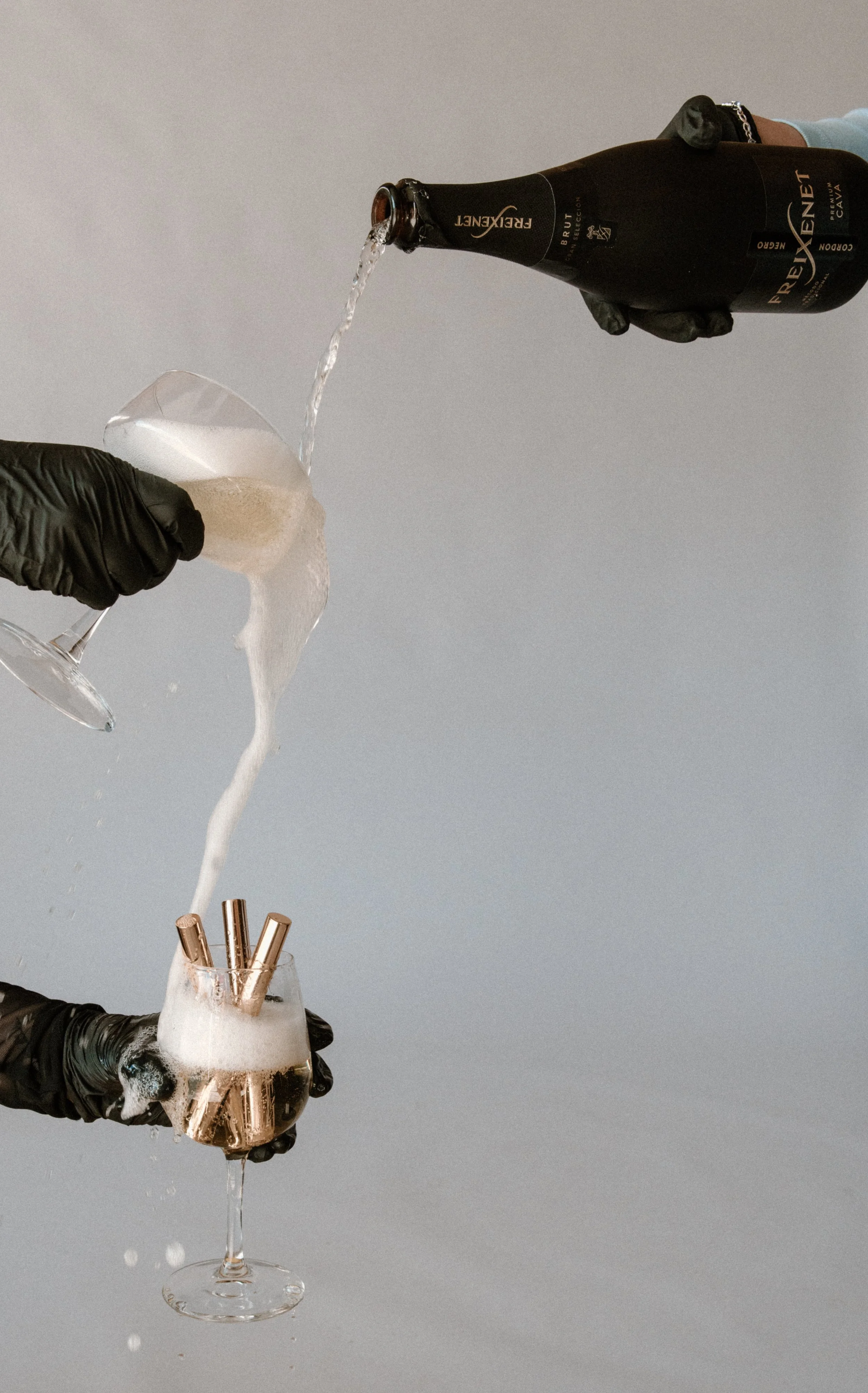 A person wearing black gloves is pouring champagne into a wine glass holding gold-colored teeth whitening pens, with foam overflowing from the glass.