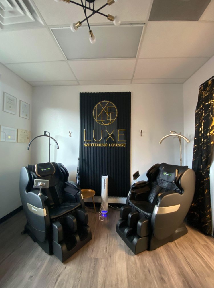 Two massage chairs facing each other in a lit room with a sign reading "LUXE Whitening Lounge" on a black textured wall in the background. The chairs have attached tablets, and there are tall, curved lamps overhead. The room has wood flooring and framed certificates on the wall.
