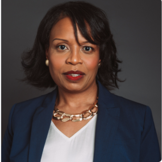Professional woman with dark hair, wearing a navy blazer, white top, and gold necklace, posing against a gray background.