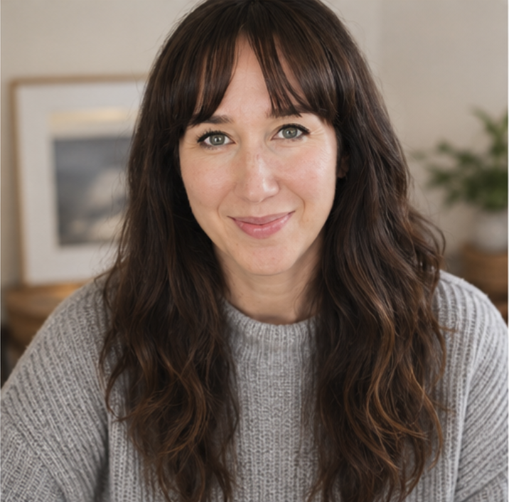 A photo of Kate the mentor with long wavy brown hair and green eyes smiling at the camera, wearing a gray sweater, indoors with a blurred background.