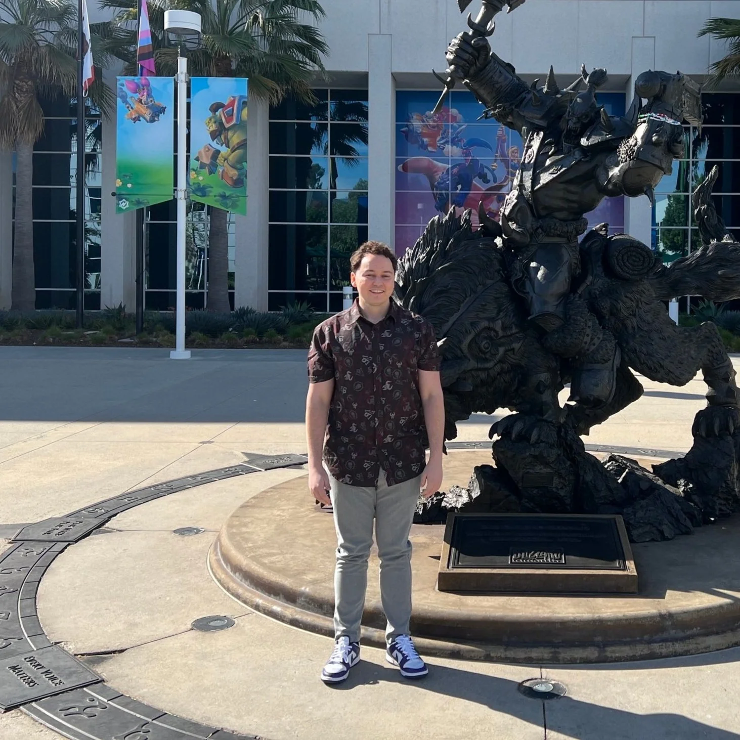 A young man standing in front of a bronze statue of a Native American warrior on a dynamic horse, outside a modern building with colorful banners and palm trees.