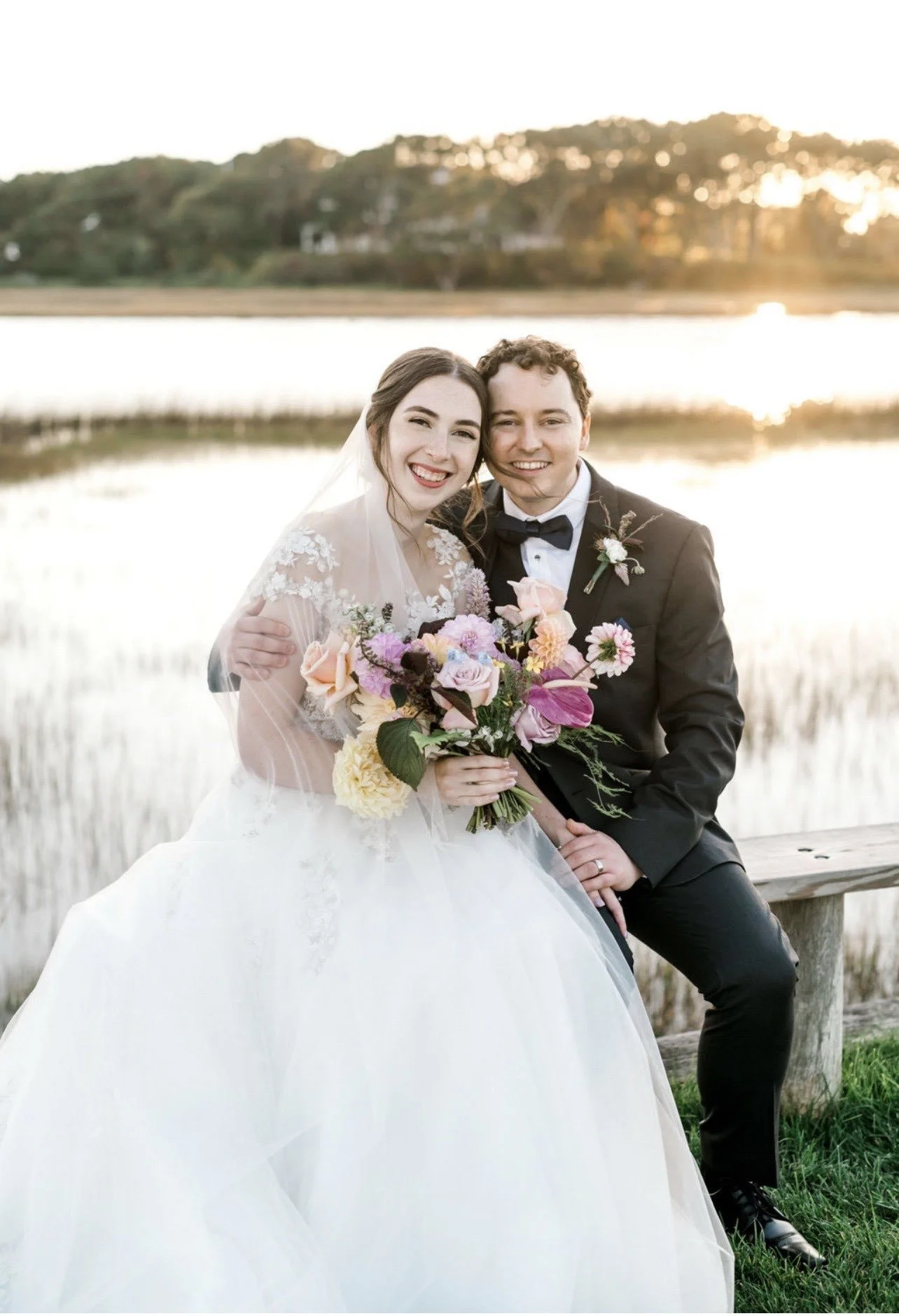 A bride and groom sitting together outdoors during sunset, smiling. The bride is wearing a white wedding dress with lace details and holding a colorful bouquet. The groom is in a black tuxedo with a bow tie. They are near a body of water with trees and houses in the background.