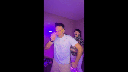 Young woman with wavy hair wearing a white camisole tops and denim shorts, standing indoors near a window with blinds, smiling at the camera.