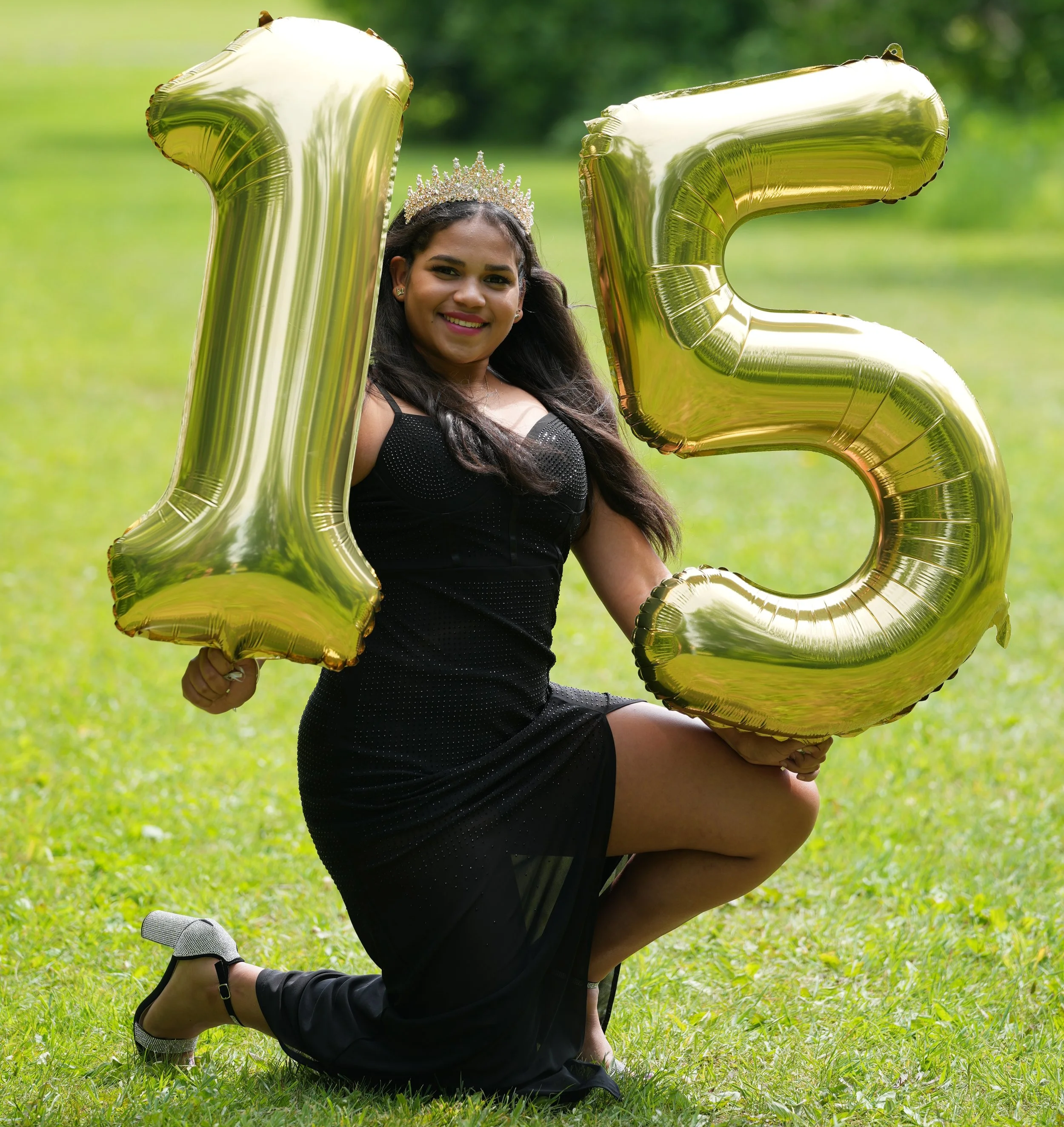 A young woman in a black dress and silver heels kneels on grass, holding large gold balloons shaped as the number 15, with a tiara on her head, celebrating her 15th birthday.