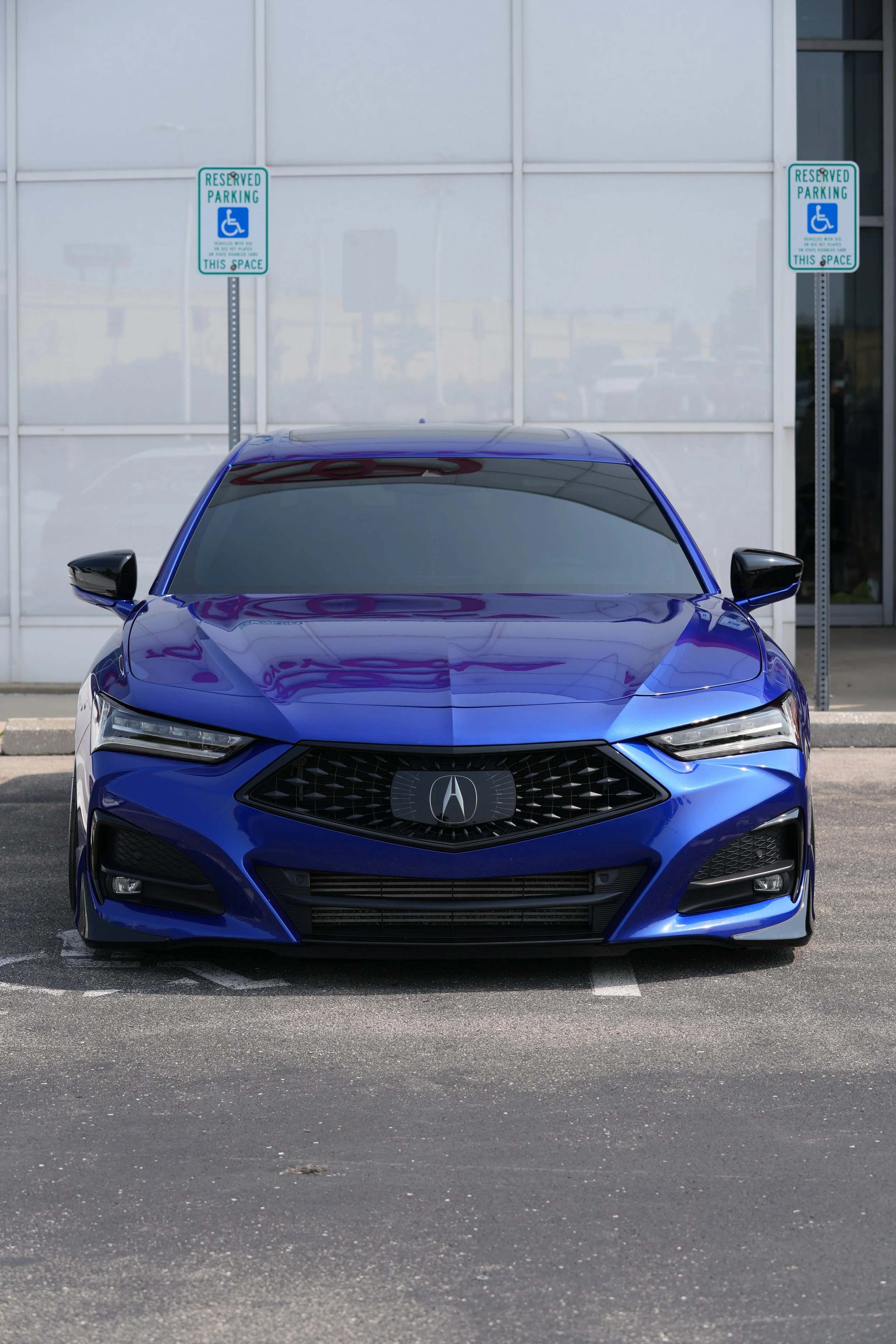 A blue Acura sedan parked in a designated handicapped parking spot next to a building with reserved parking signs for handicapped individuals.