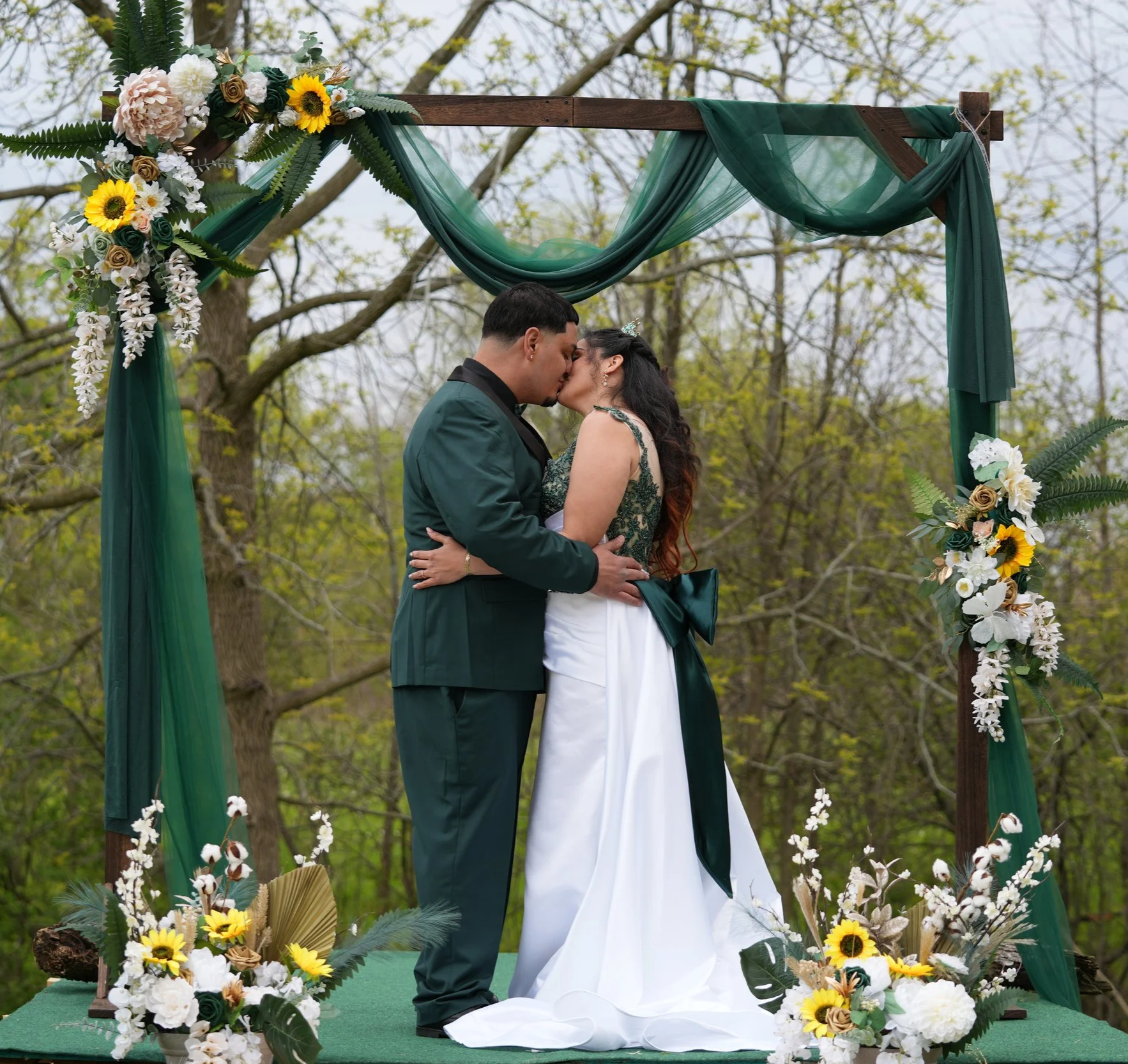A couple kissing at their outdoor wedding ceremony under a decorated wooden arch with green fabric and floral arrangements, surrounded by trees.