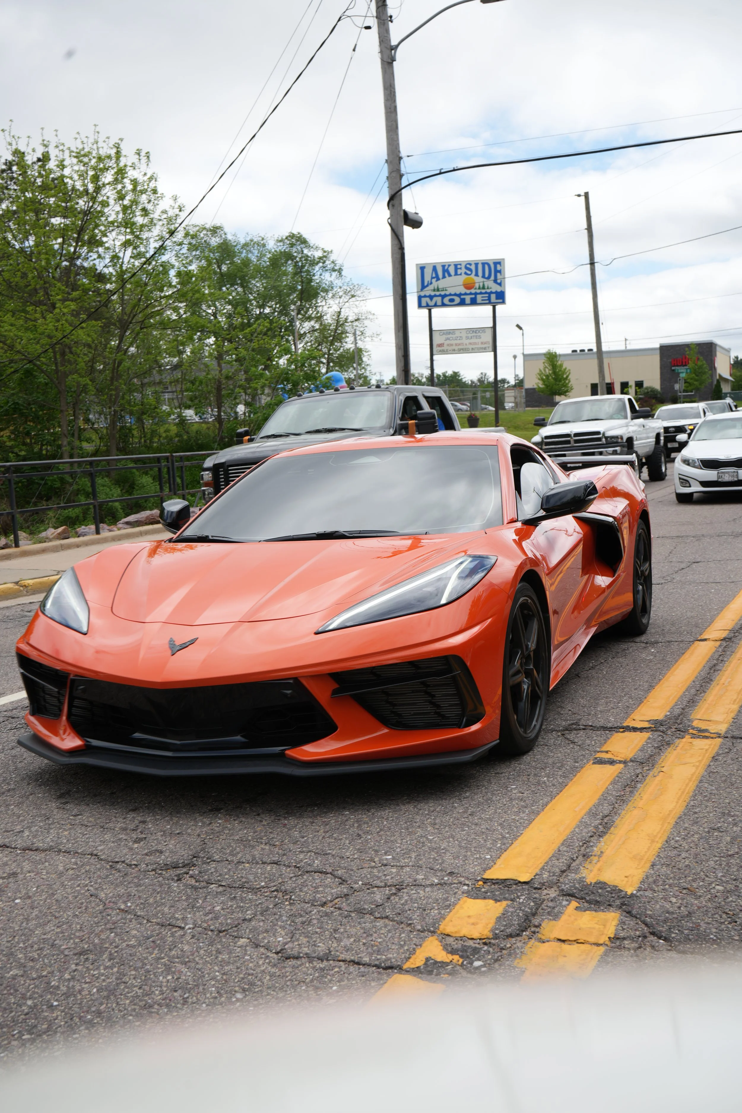 An orange Chevrolet Corvette sports car parked on the street near a yellow line, with other vehicles and a motel sign in the background.