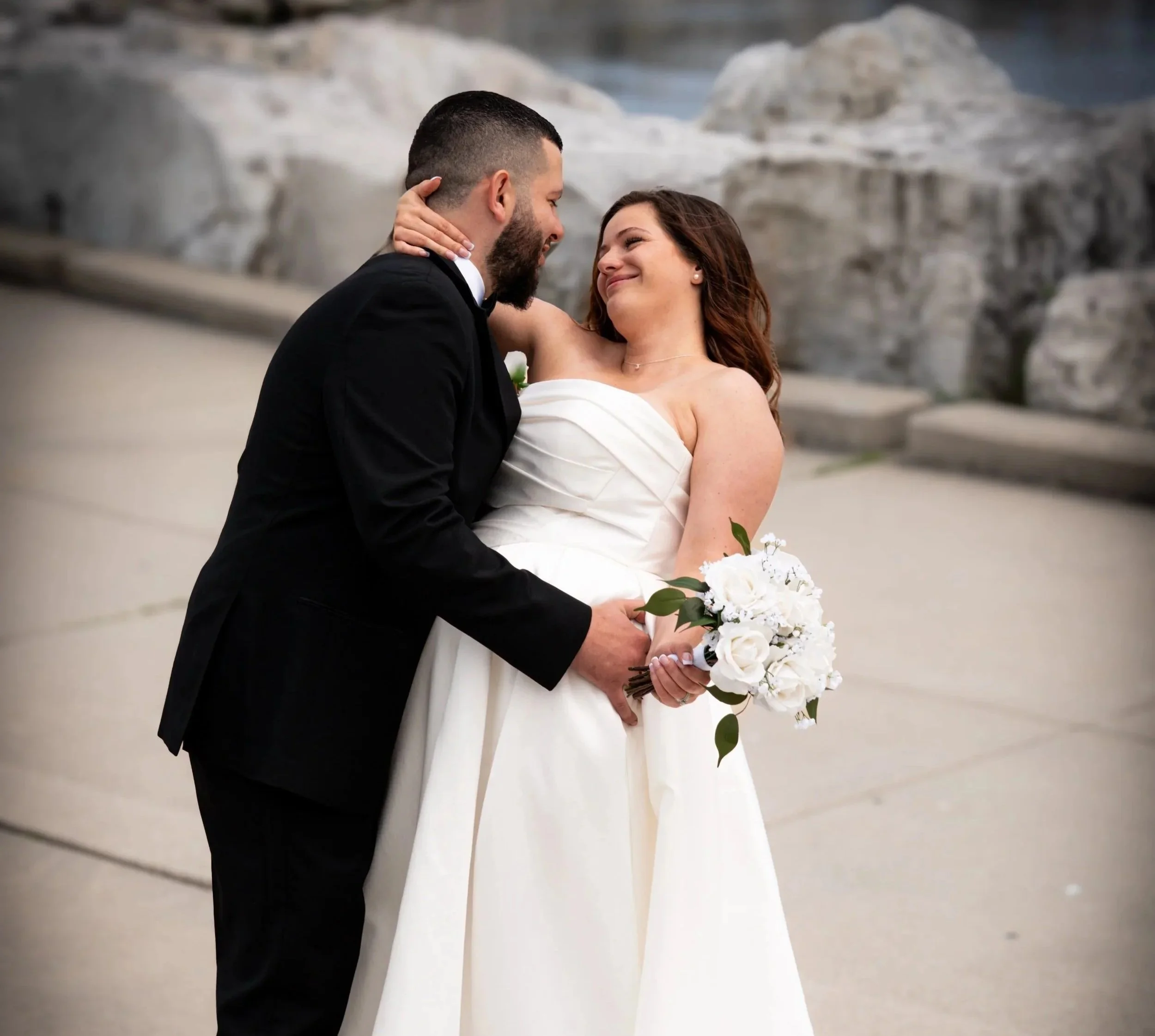 A newlywed couple sharing an intimate moment on their wedding day, with the bride holding a bouquet of white flowers, and the groom dressed in a black suit, set against a rocky outdoor background.