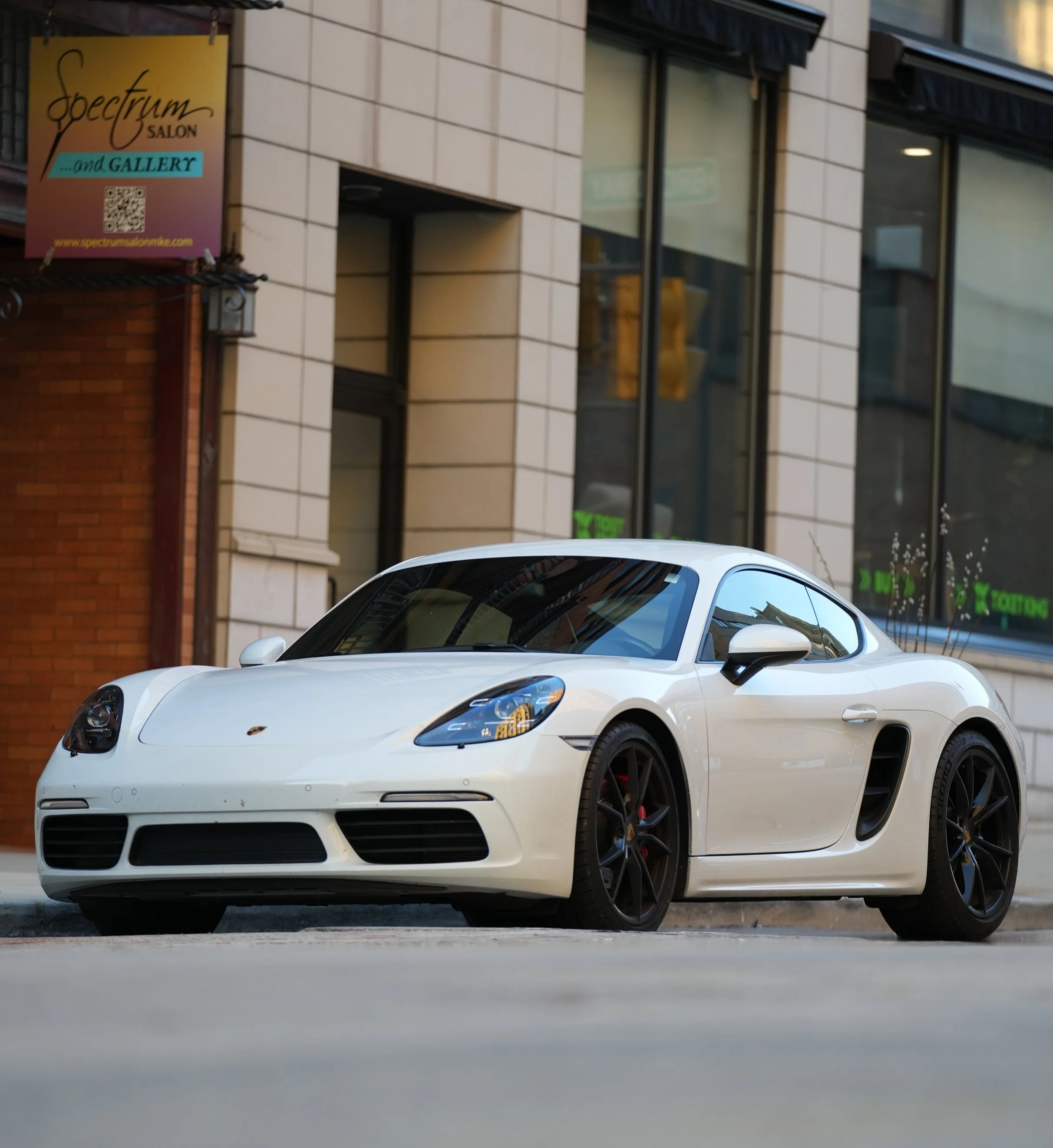 A white sports car parked on a city street in front of a building with large windows and a sign for Spectrum Salon.