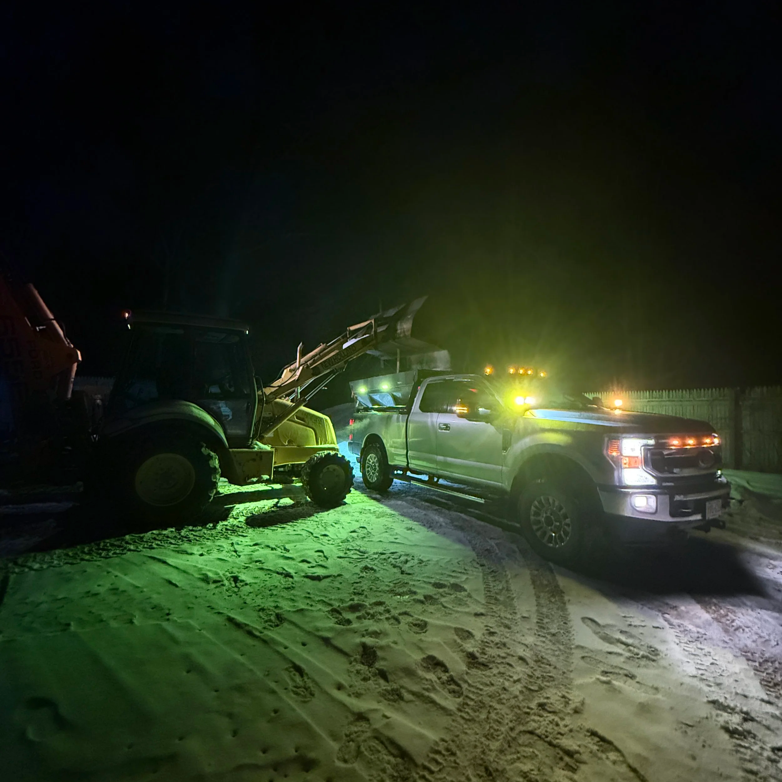 Nighttime scene of construction vehicles on a sandy construction site, illuminated by green and yellow lights.