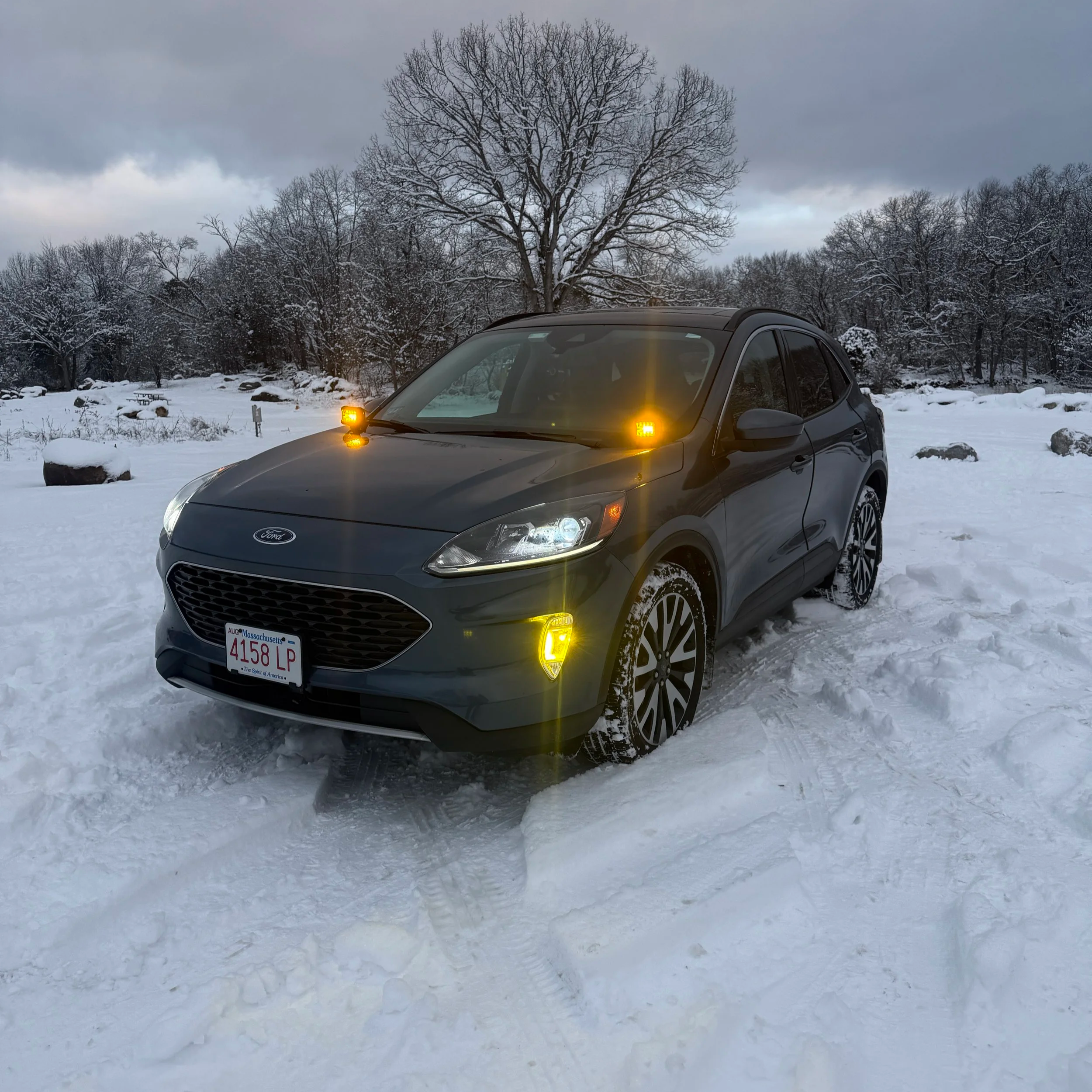 A gray car with orange warning lights on a snowy landscape with snow-covered trees and a cloudy sky.