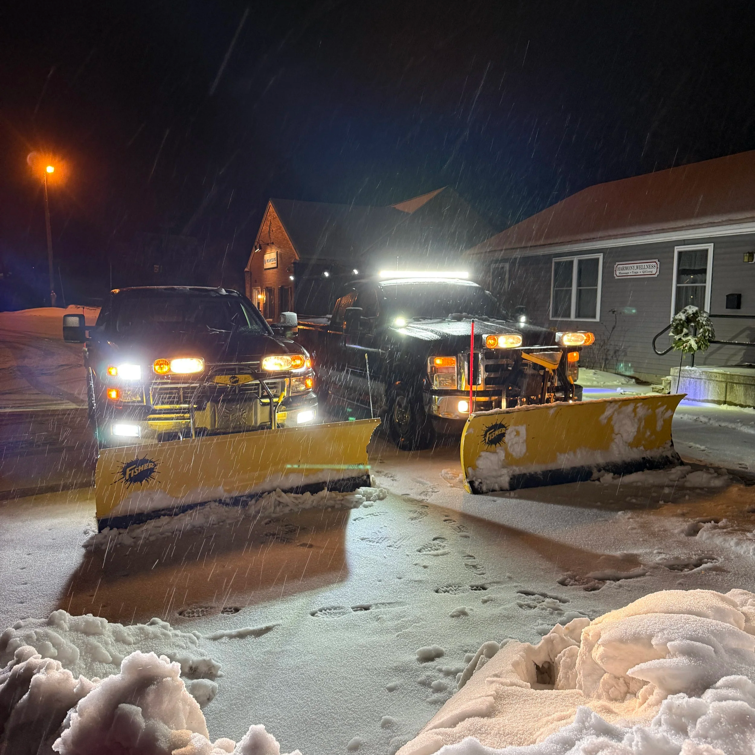 Nighttime scene with snow-covered ground, several parked cars, snowplow visible, building with sign reading 'HARMONY WELLNESS', snow falling, street lamp illuminating the scene.