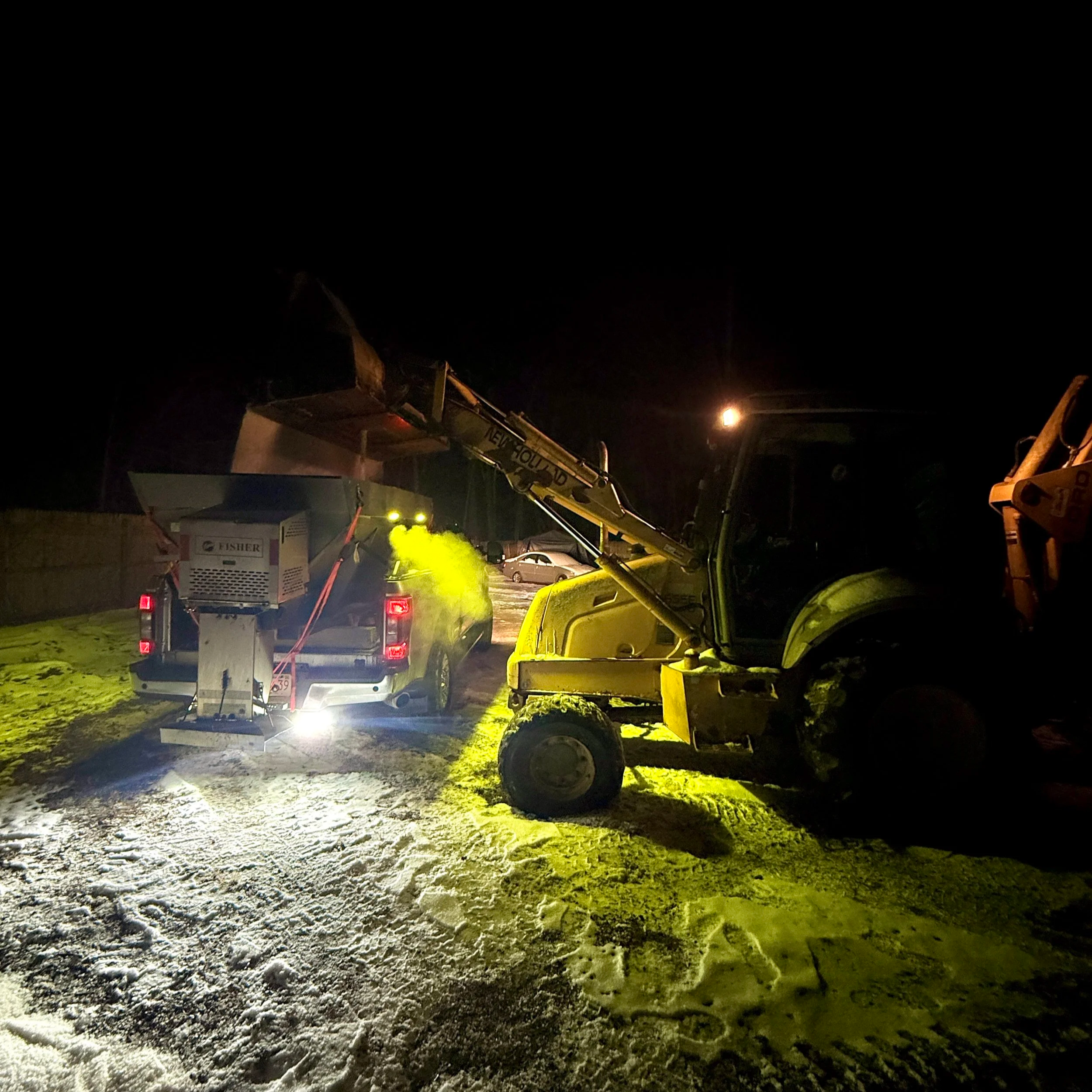 Nighttime construction scene with trucks, equipment, and lights illuminating the area with snow patches on the ground.