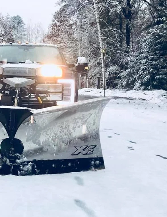 A snow-covered road with a snowplow clearing snow, the plow's blade in front of a vehicle with its headlights on, and snow-covered trees lining the road.