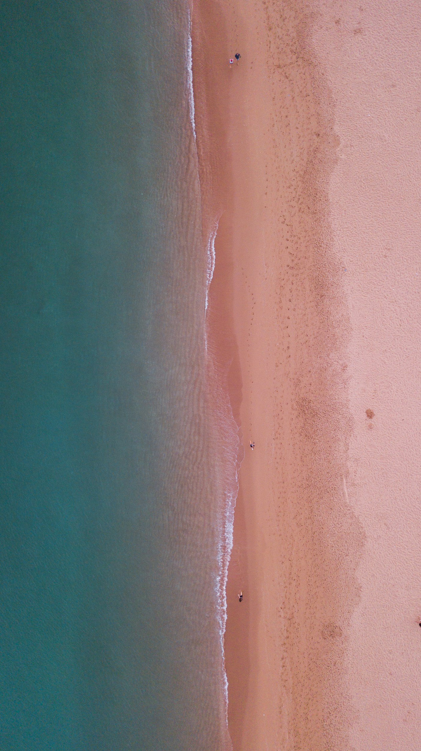 Aerial view of a beach with pinkish sand, gentle waves coming ashore, and a few people relaxing under umbrellas.