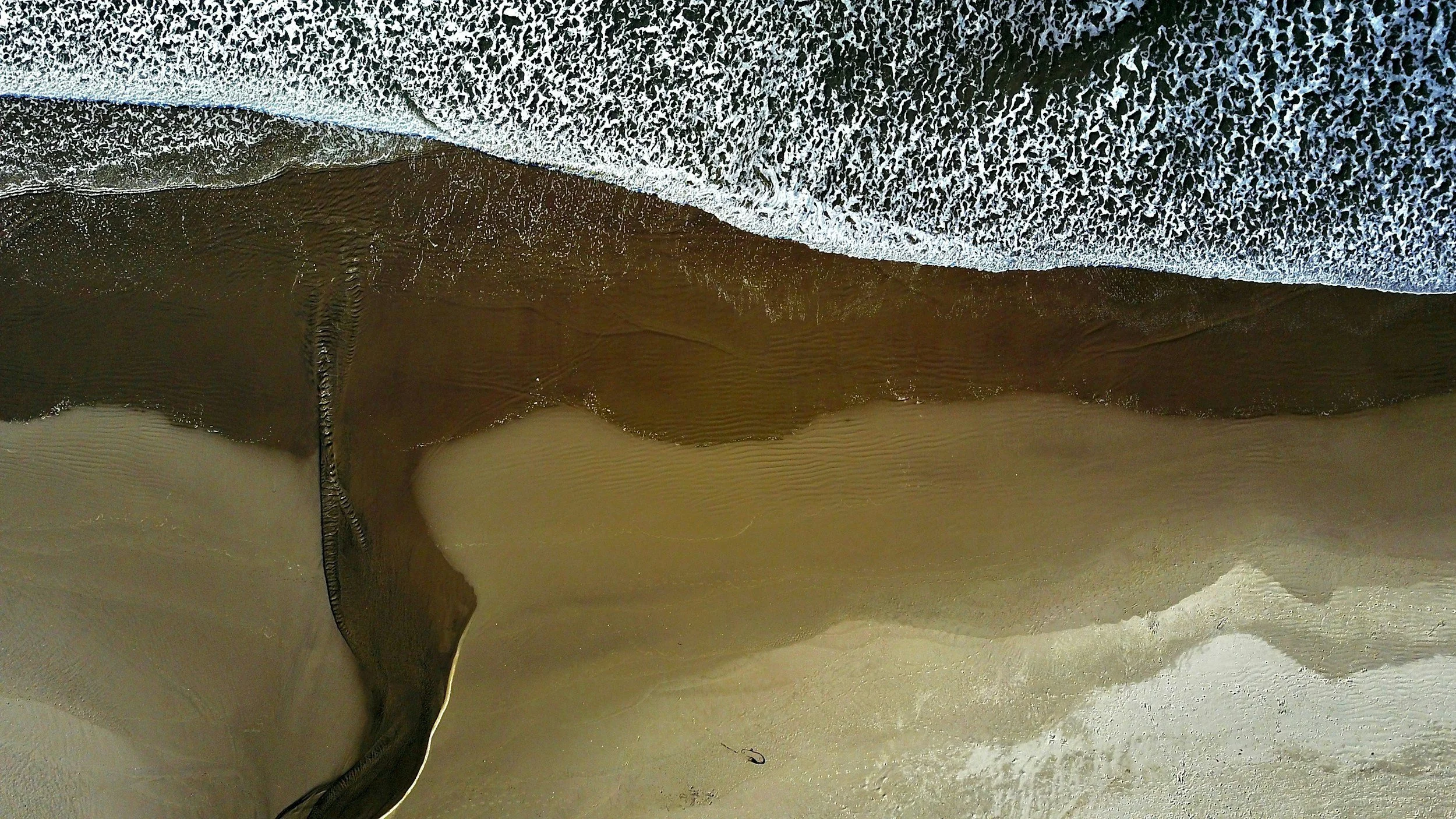 An aerial view of a beach showing the shoreline with waves, wet sand, and a small black object on the sand.