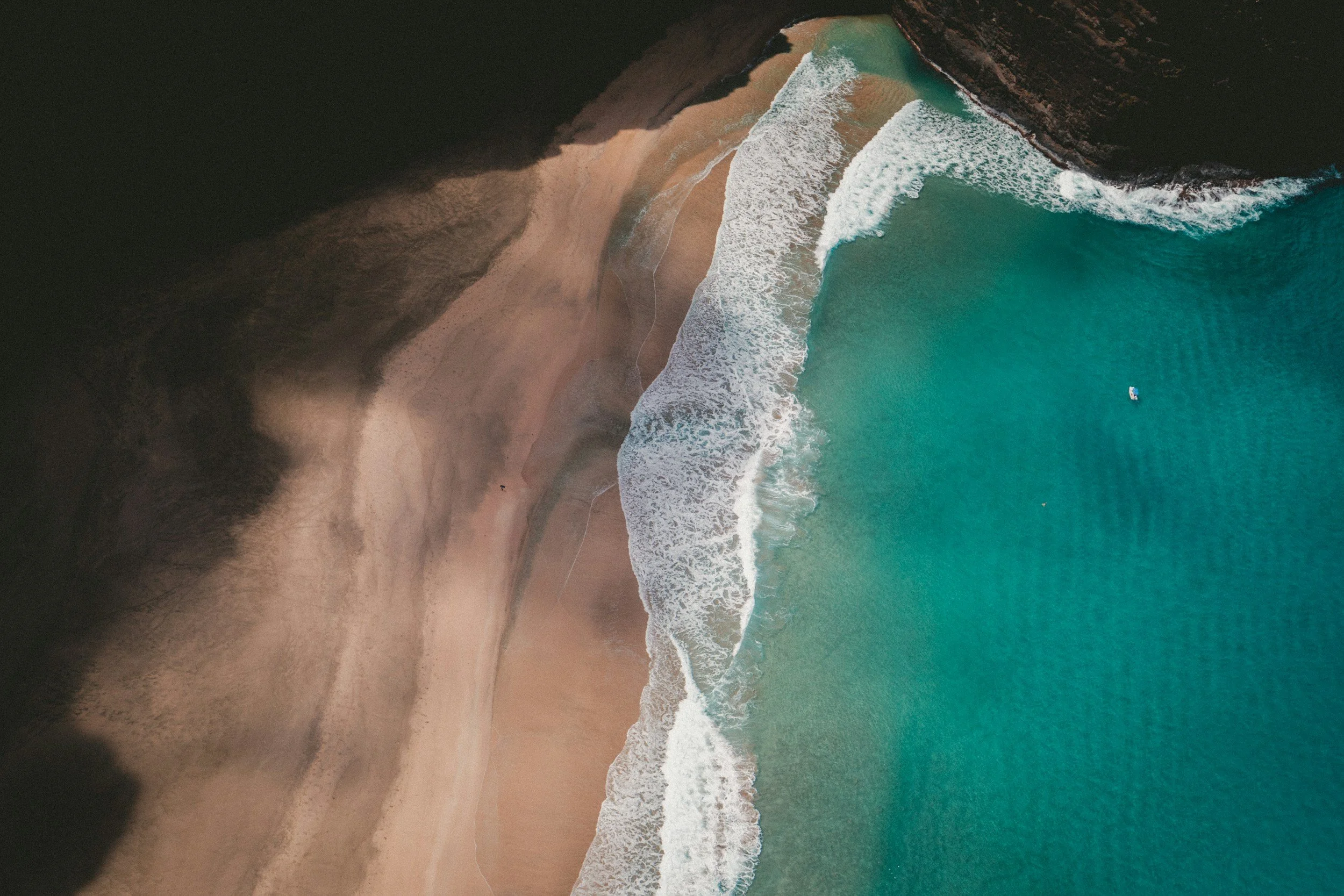Aerial view of a beach with turquoise water, white waves, sandy shore, and dark cliffs.