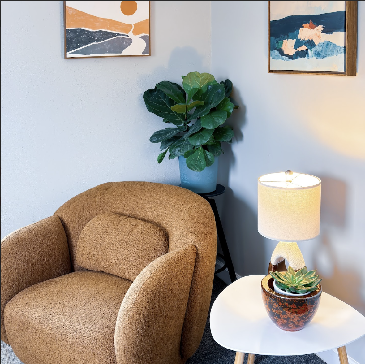 Cozy living room corner with tan upholstered armchair, white side table with a potted succulent, a table lamp, a large potted plant on a black stand, and abstract artwork on white walls.