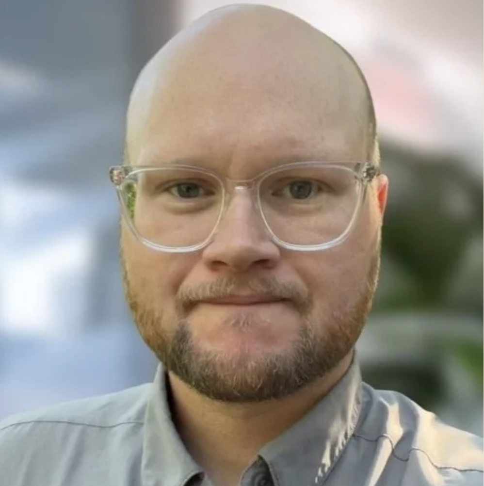 Close-up of a man with glasses and a beard, wearing a collared shirt, outdoors.