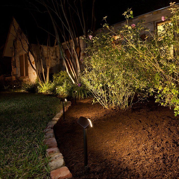 Nighttime garden with pathway lights illuminating plants and trees along a stone-edged garden bed.