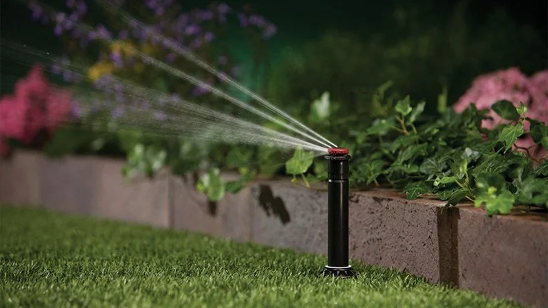 A garden sprinkler spraying water over grass and flowers at night.