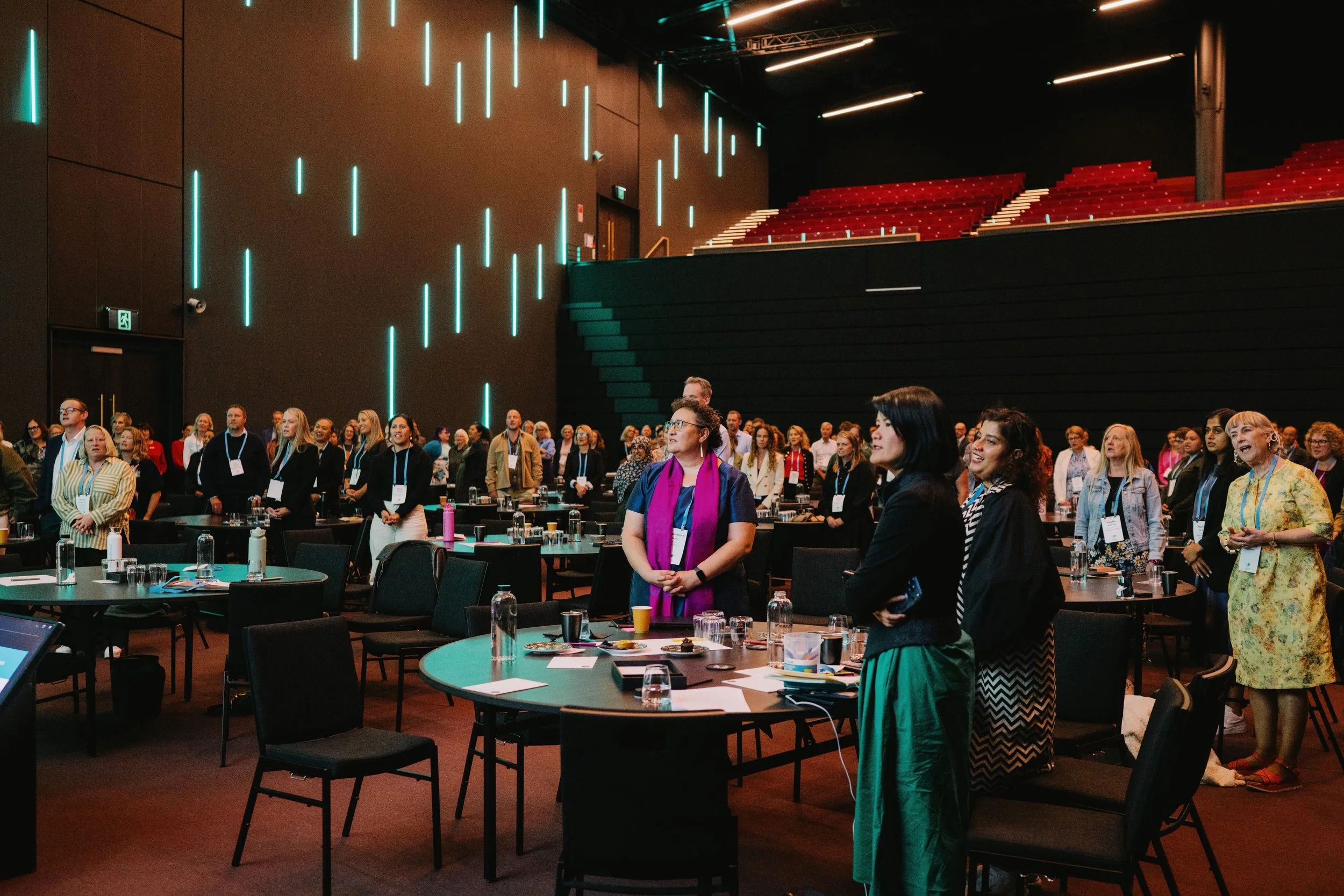 Delegates join waiata in the main theatre. Shows people standing, wearing their conference lanyards and looking hard left of camera.