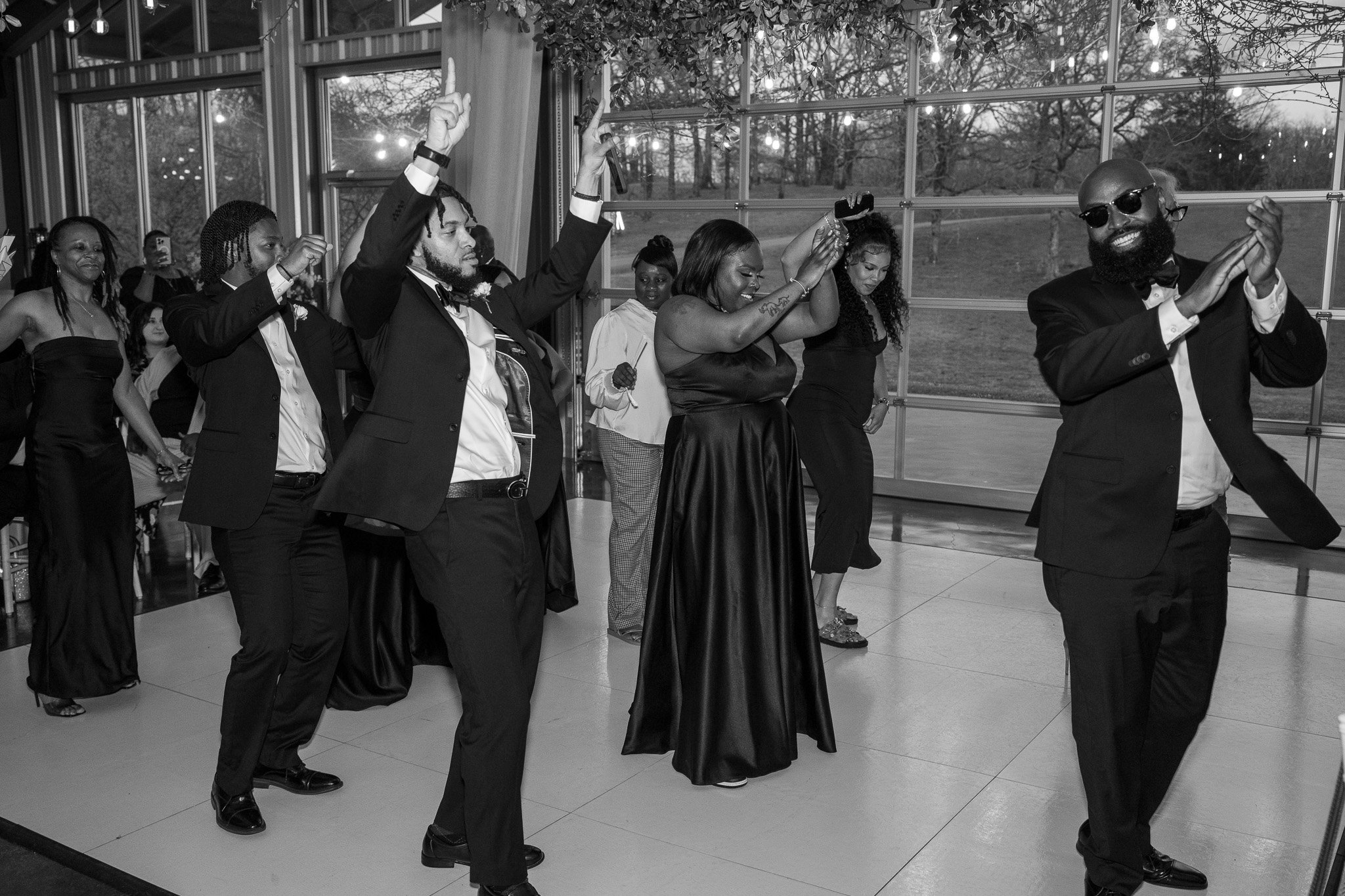 Group of people in formal attire dancing at an indoor event, with men in suits and women in dresses, in a room with large windows and greenery.