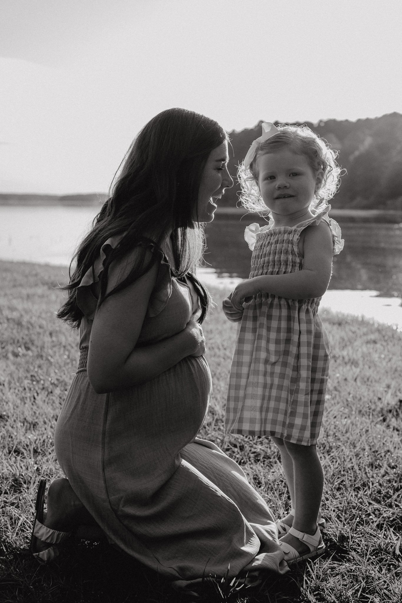 A pregnant woman kneeling on grass, smiling and interacting with a young girl near a lake, in black and white.