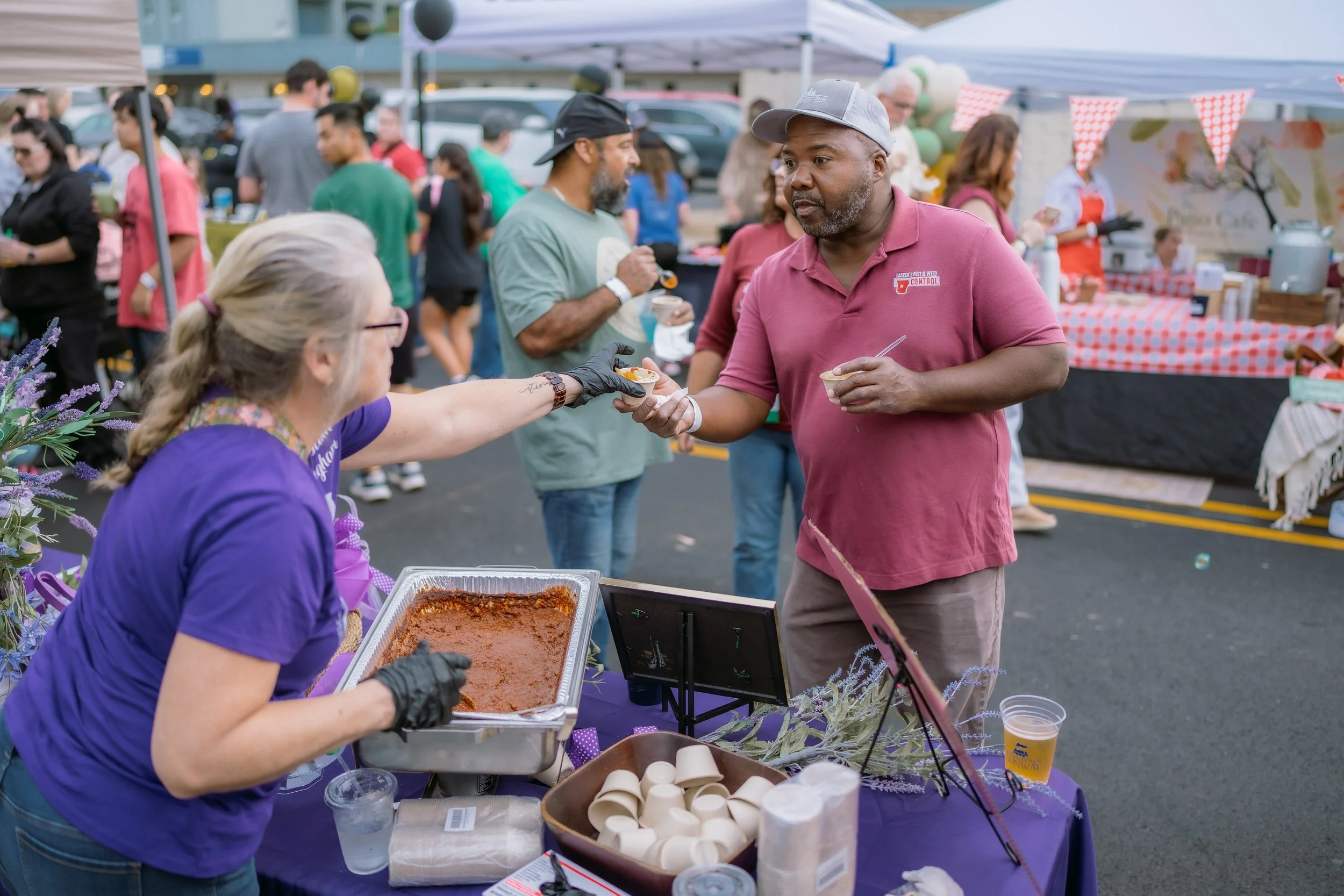 A woman in a purple shirt hands a small cup of food to a man in a pink shirt at an outdoor food stall during a community festival.
