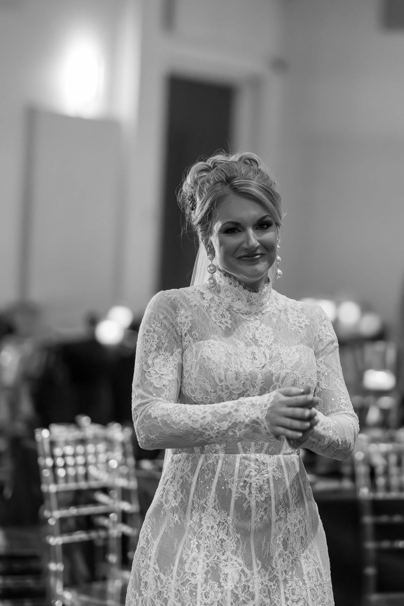 Bride in lace wedding dress smiling indoors, wearing large earrings.