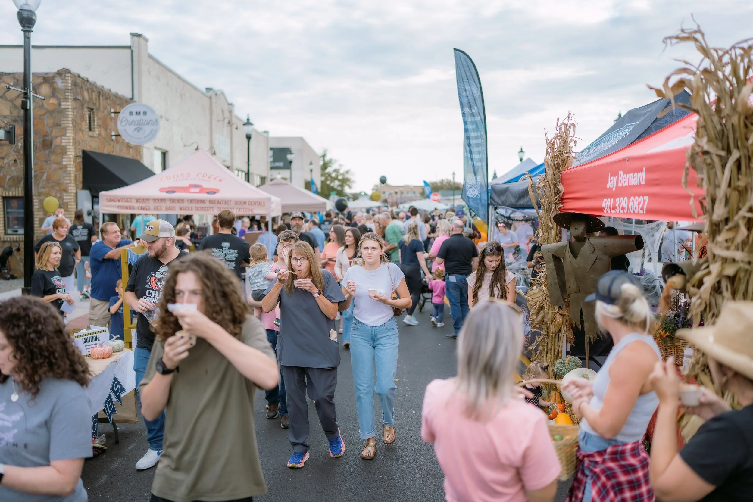 People walking at an outdoor fall festival or market with vendor stalls, pumpkin displays, and tents under a cloudy sky.