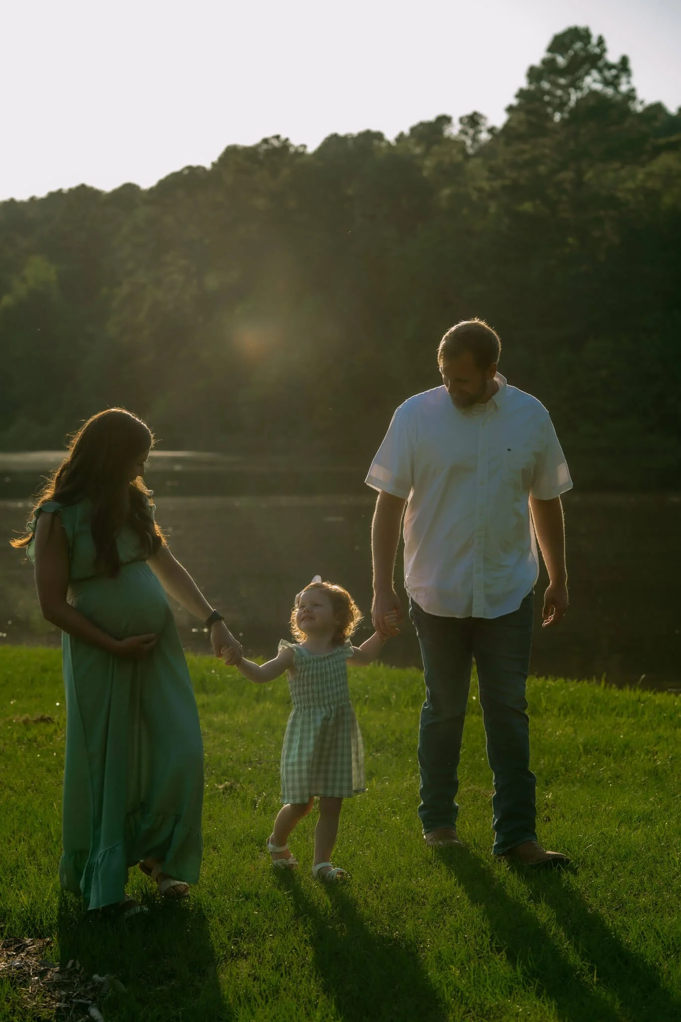 A family walking hand in hand by a lake during sunset, with a pregnant woman, a man, and a little girl.