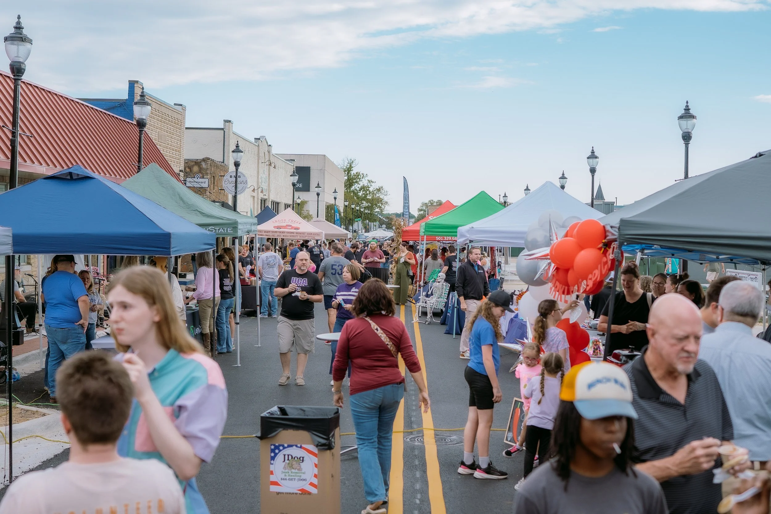 People attending outdoor street fair with vendor booths and balloons on a closed road.
