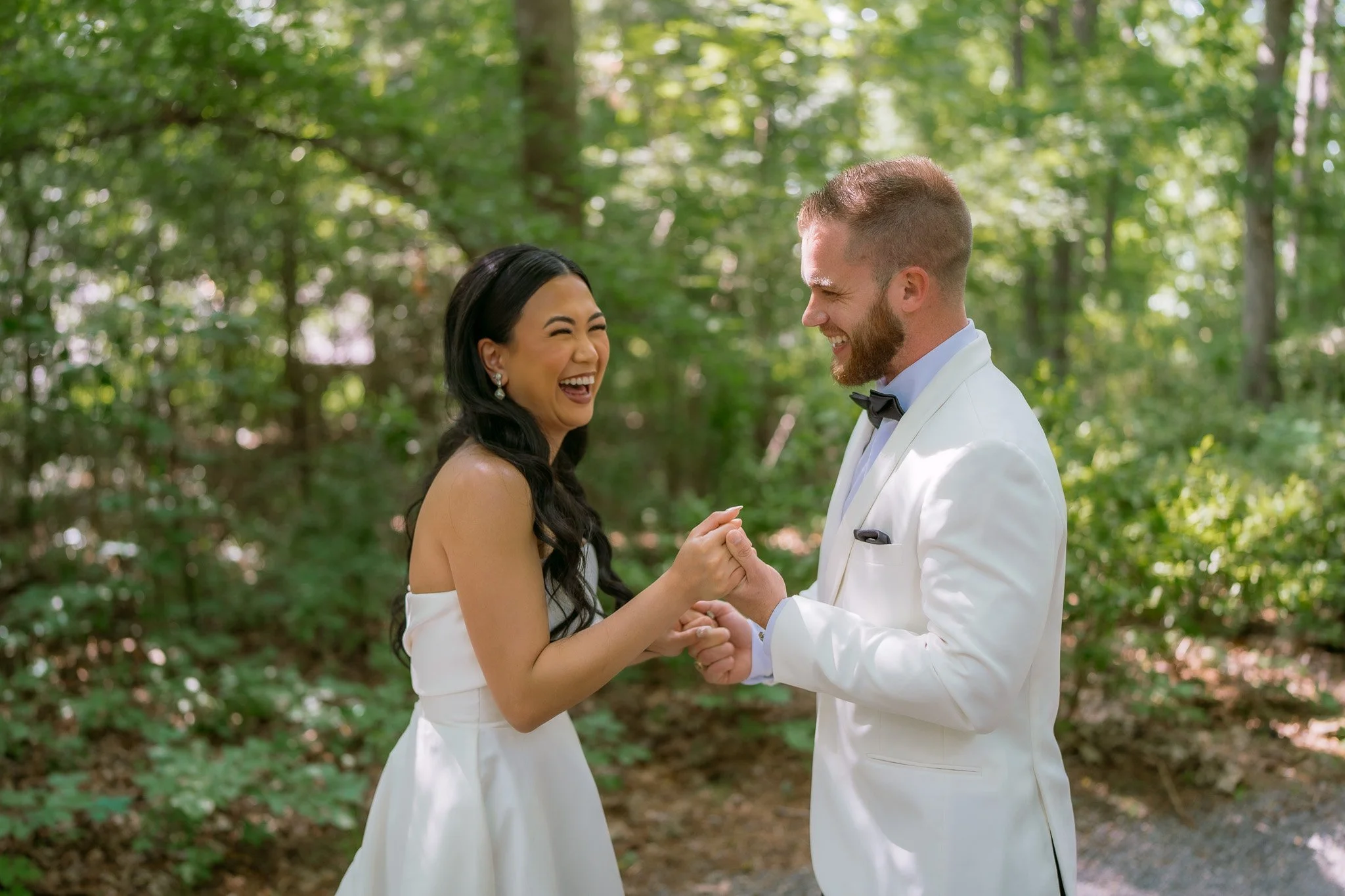 A bride and groom holding hands and smiling at each other outdoors in a wooded area during daytime.