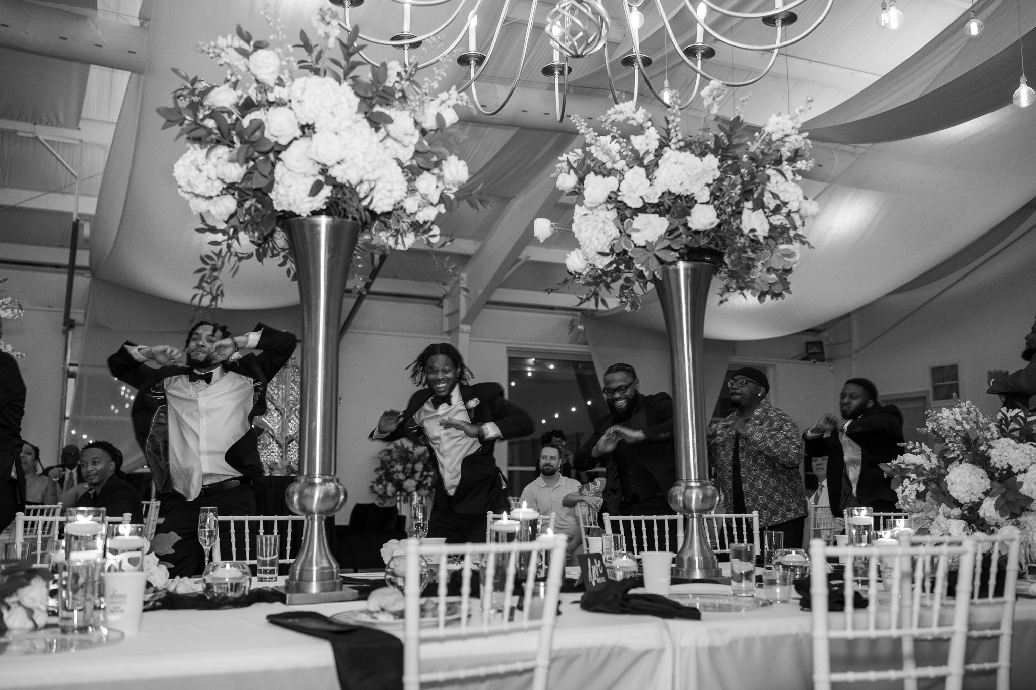 Black and white photo of people dancing in a decorated event space with floral arrangements and chandeliers. Table settings and chairs visible in the foreground.