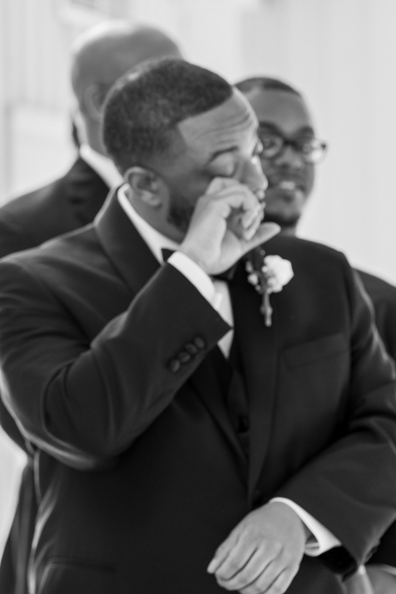 Man in a tuxedo wiping tears at a wedding, with another man smiling in the background.