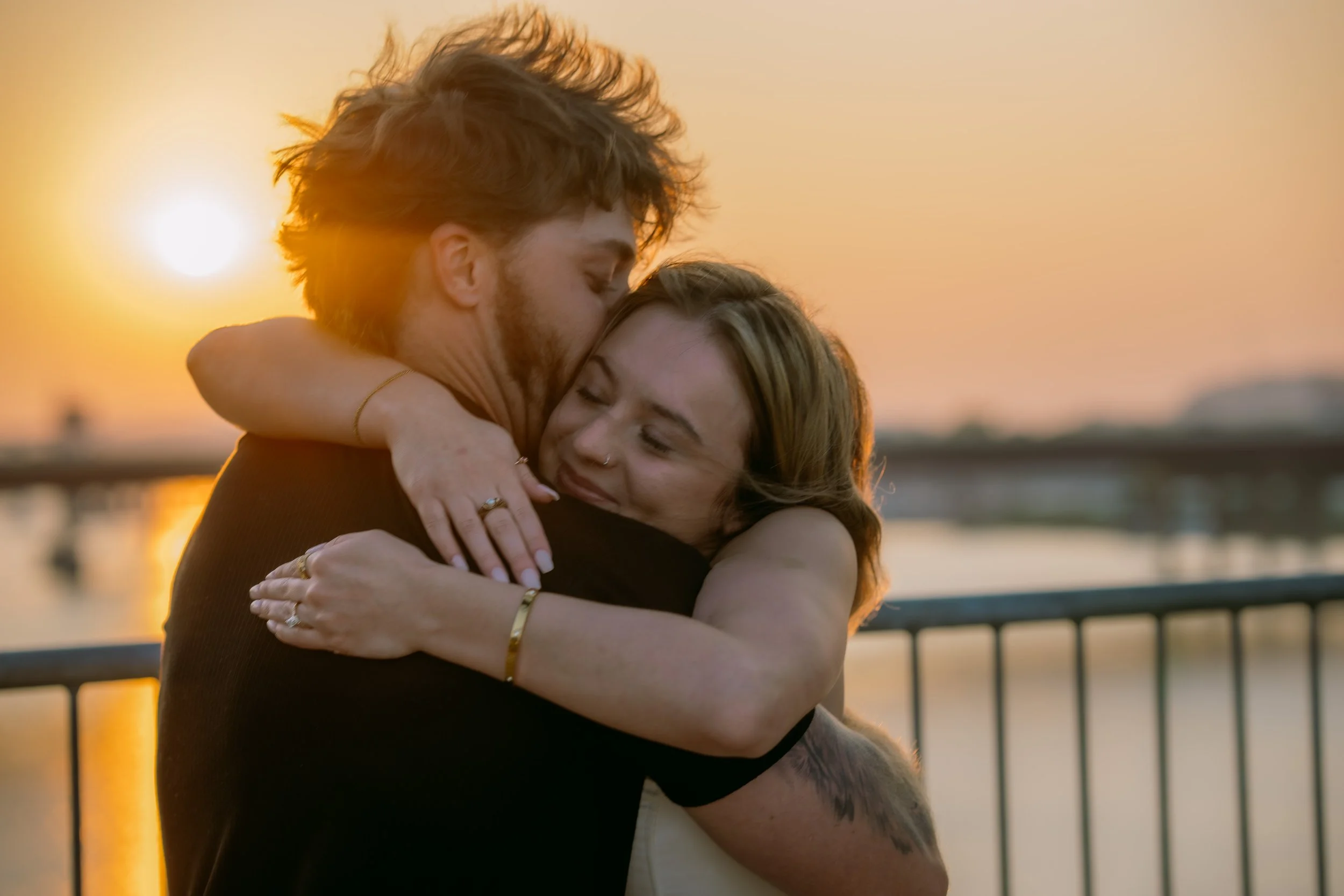 A couple embracing during sunset near a body of water with a bridge in the background.