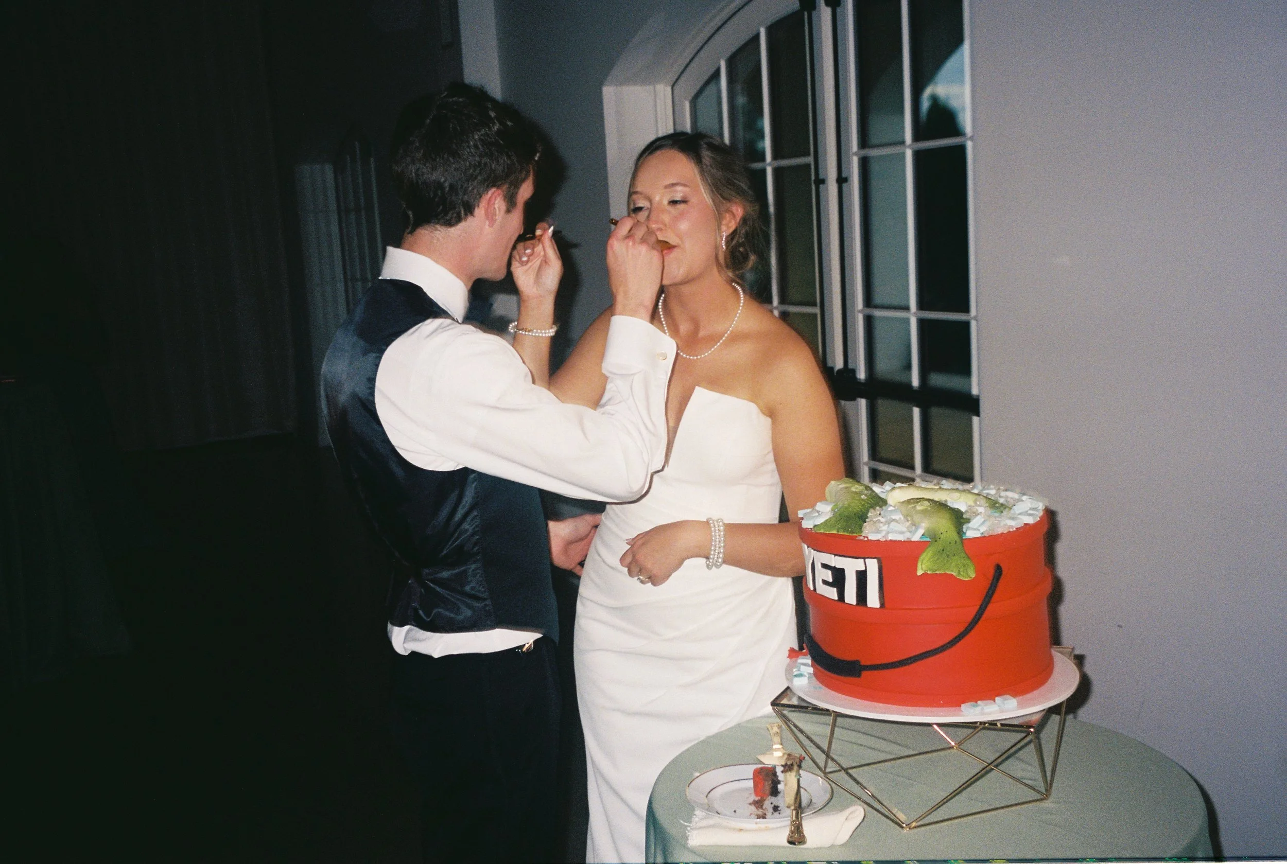 A bride and groom at their wedding reception, with the groom feeding the bride cake. There is a wedding cake multicolored with a smiley face on a table nearby.